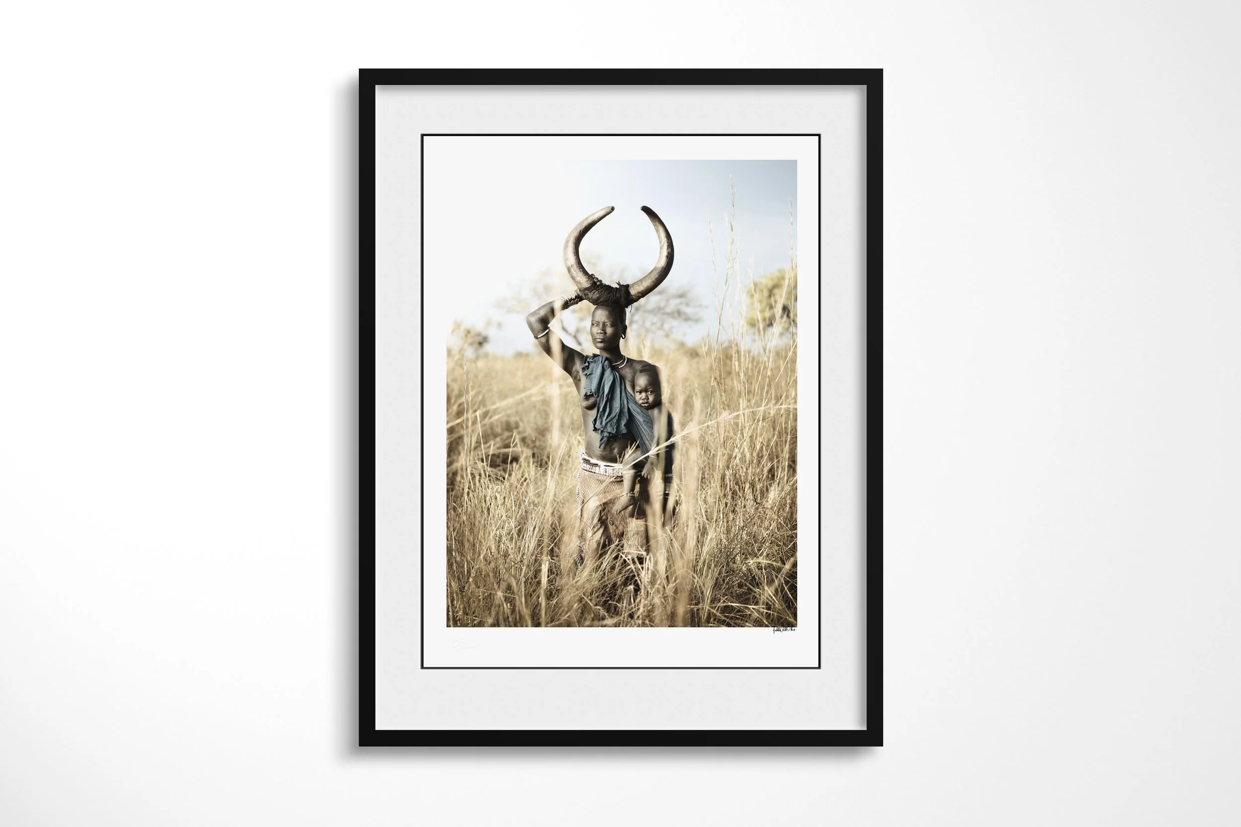 Framed presentation of Markori & Idho in a black frame and white mount, showing a Mursi woman carrying a child beneath a large curved-horn headdress in tall grass.