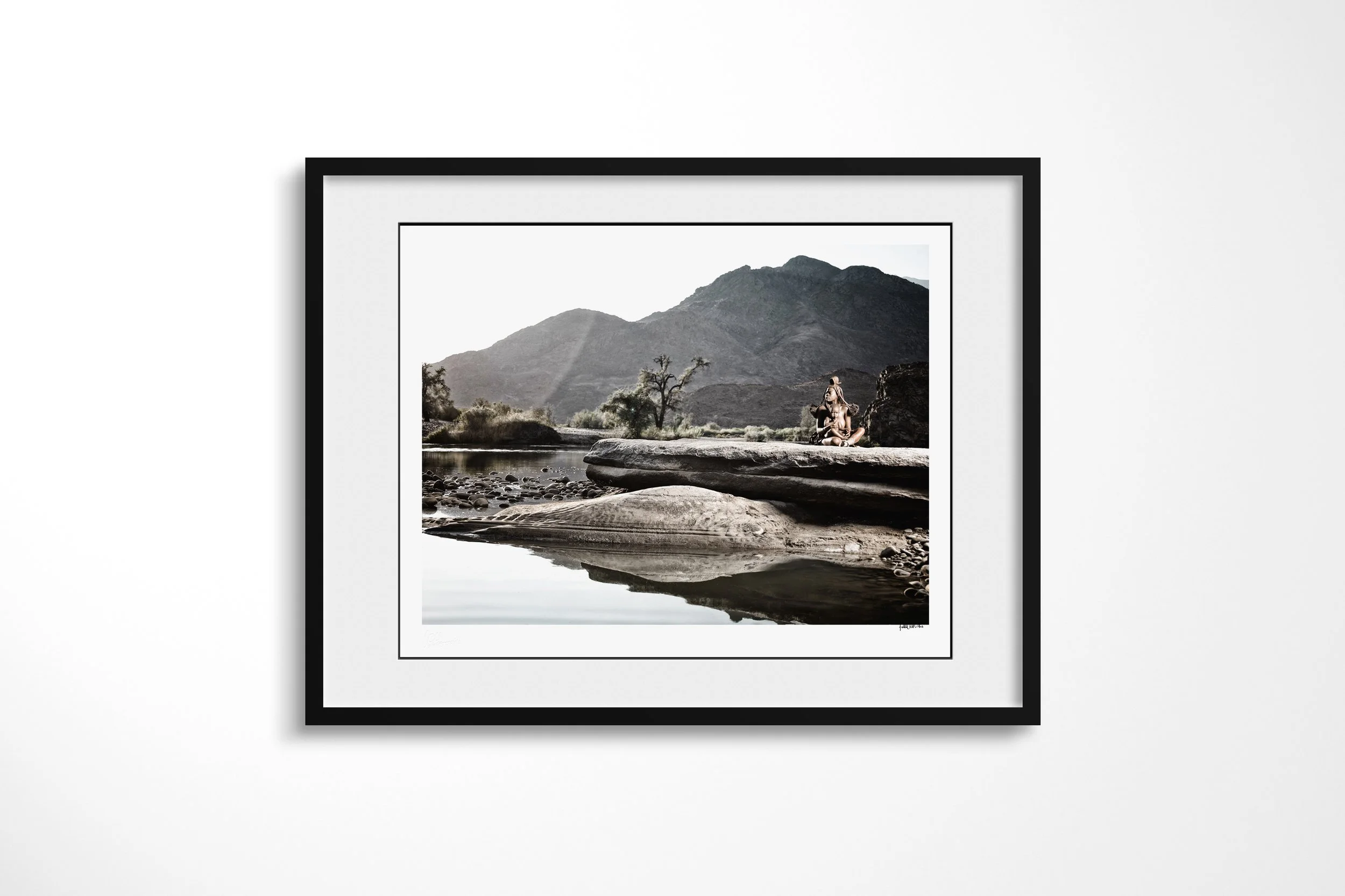 Framed presentation of Tjikuria in a black frame and white mount, showing Tjikuria and a child seated on a broad rock beside the Kunene River beneath dark mountains.