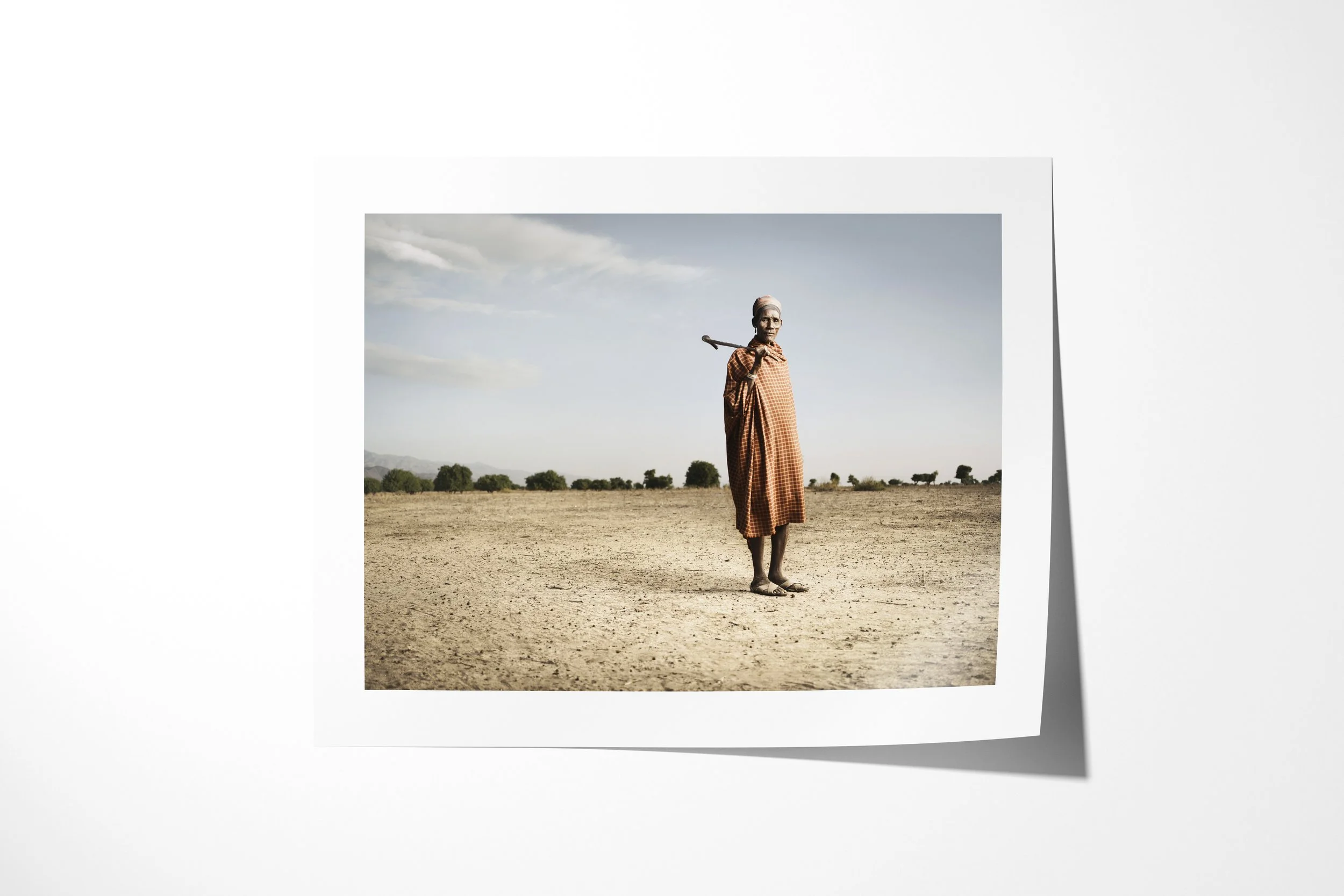 Fine art paper print of Aregore, showing an older Arbore man wrapped in checked cloth standing with a staff across his shoulders on open ground.