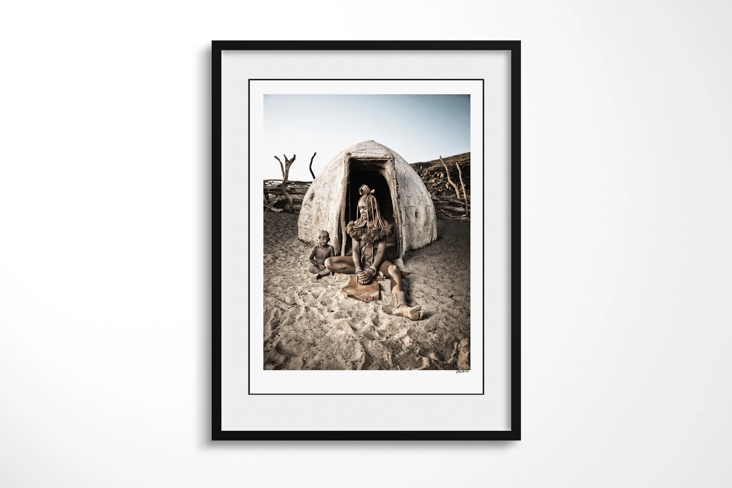 Framed presentation of Ngaringombe And Her Son in a black frame and white mount, showing Ngaringombe seated with her son outside a domed Himba dwelling while grinding ochre on a stone.