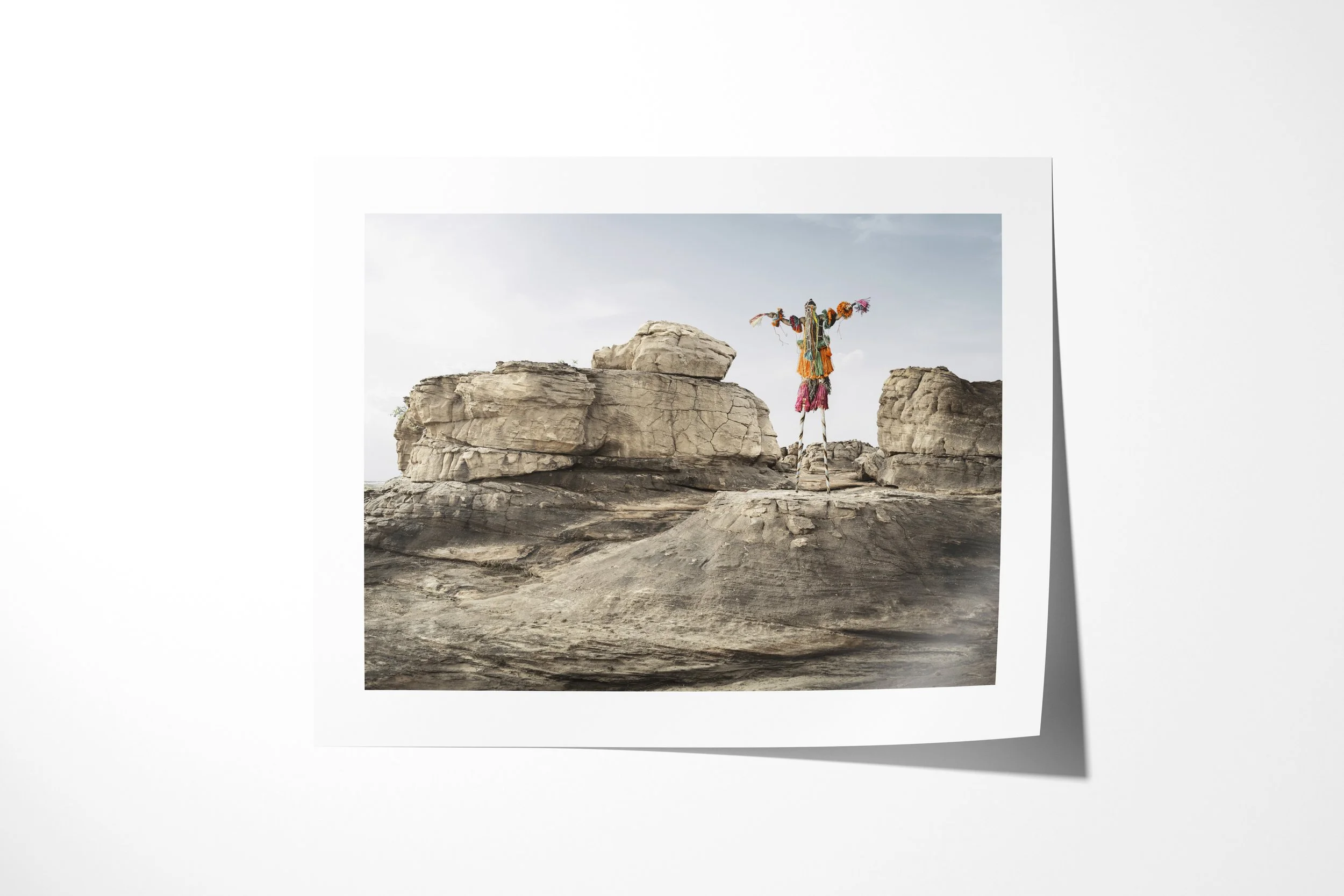 Fine art paper print of Tegué Tanga (Mask), showing a Dogon stilt mask standing between broad sandstone outcrops.