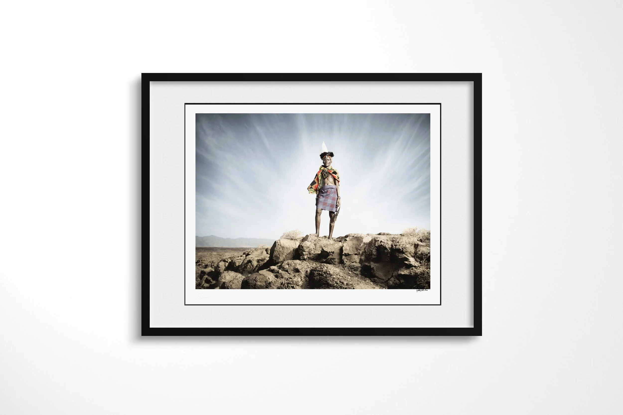 Framed presentation of Ayapan in a black frame and white mount, showing Ayapan standing on dark rocks in a feathered headdress beneath a streaked sky.