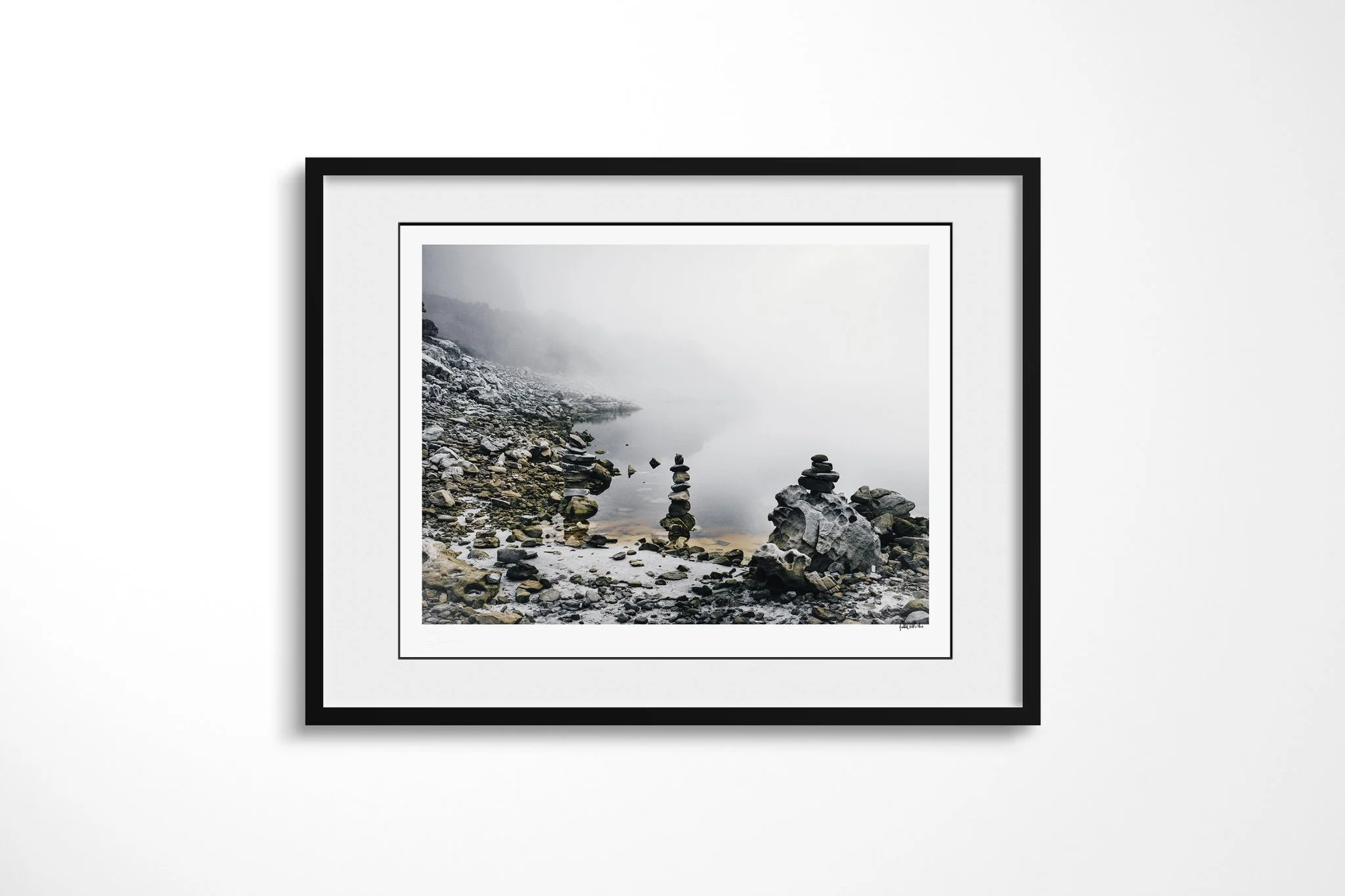 Framed fine art presentation of Cairn in a black frame with a white mount, depicting balanced stone cairns beside still water on a rocky shore fading into pale fog.