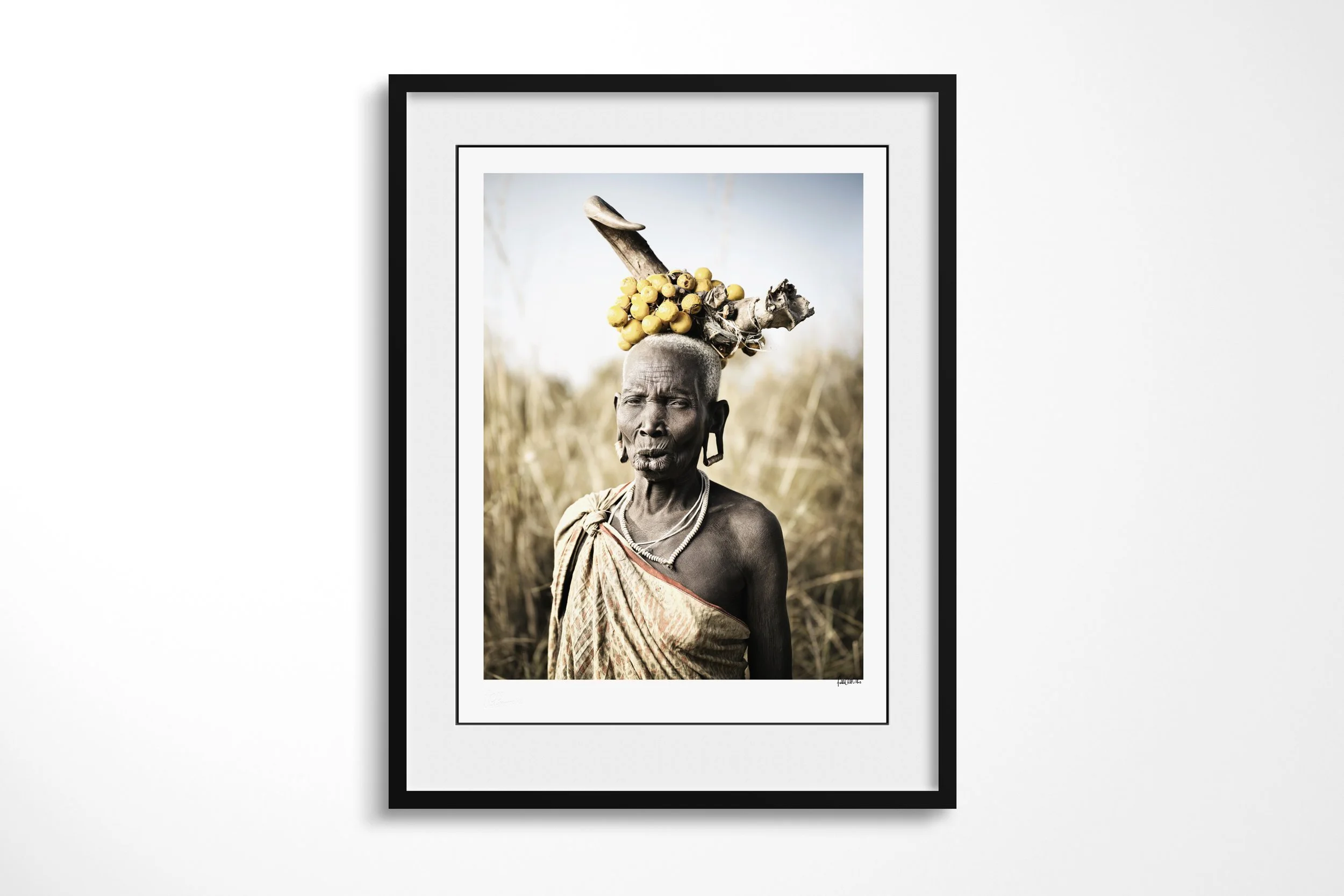 Framed presentation of Wechidi in a black frame and white mount, showing an older Mursi woman with elongated lip and earlobes wearing a horn-and-fruit headdress in tall grass.
