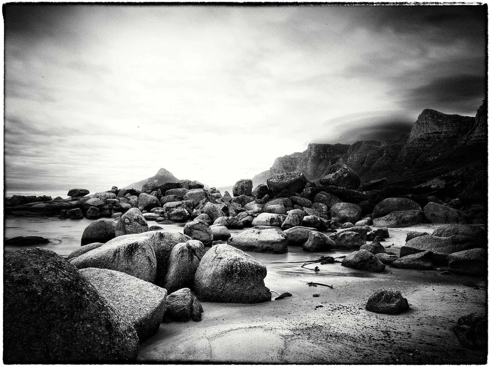 Black-and-white boulder beach beneath Cape Town's Twelve Apostles cliffs, with low cloud and softened seawater.