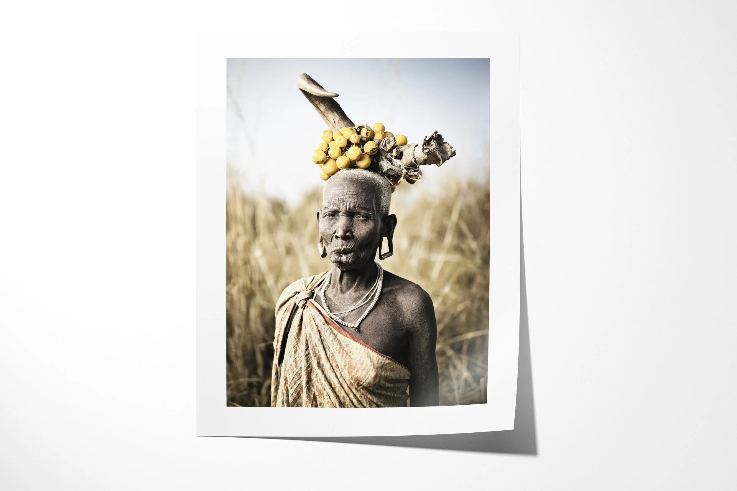 Fine art paper print of Wechidi, showing an older Mursi woman with elongated lip and earlobes wearing a horn-and-fruit headdress in tall grass.
