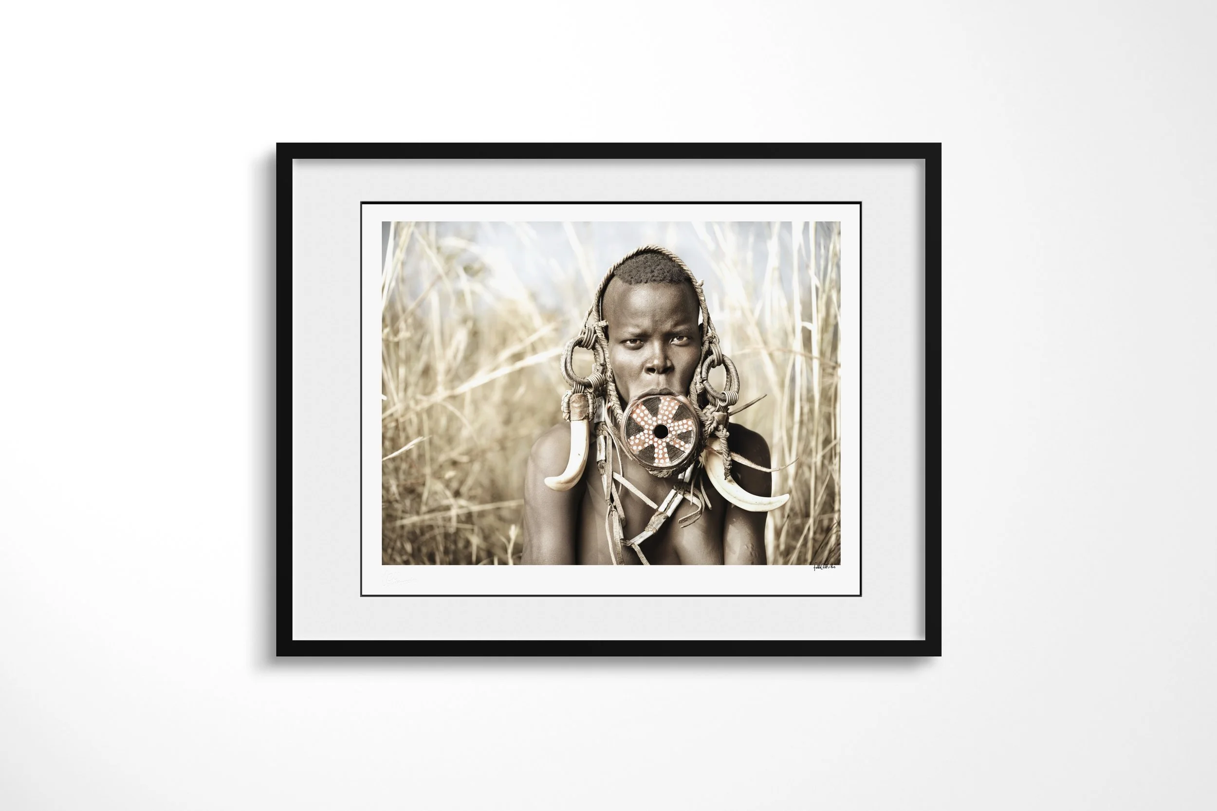 Framed presentation of Uluio in a black frame and white mount, showing a close portrait of a Mursi woman wearing a painted lip plate, large tusks and layered ornaments.