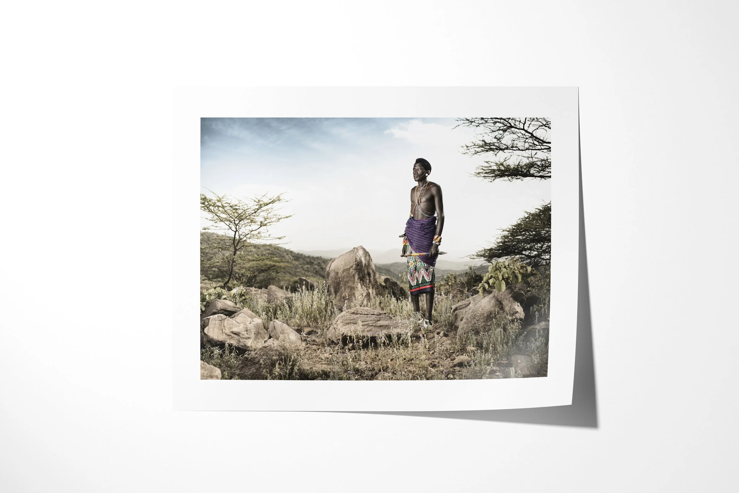 Fine art paper print of Lkatinga, showing a Samburu man in purple and patterned clothing standing among rocks in the Matthews Mountains.