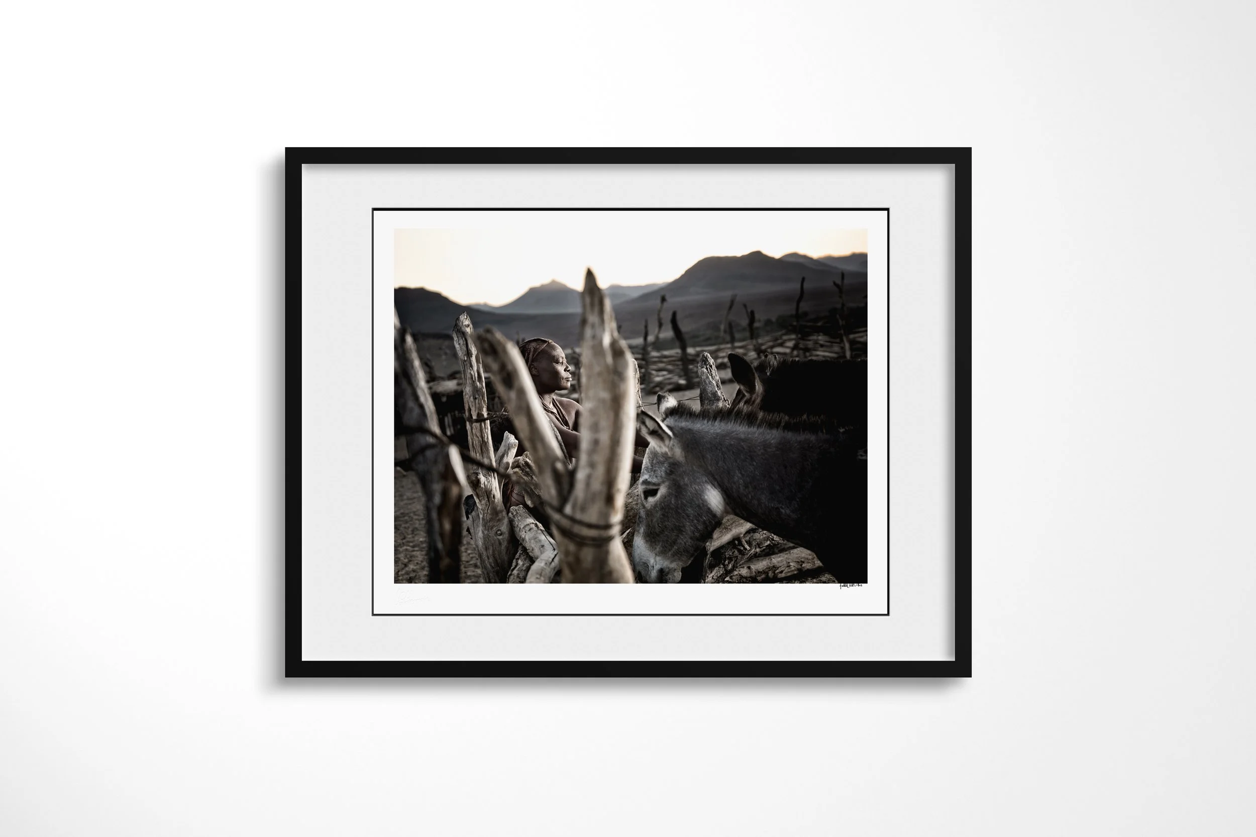 Framed presentation of Tjihaumwa in a black frame and white mount, showing Tjihaumwa seated among cattle horns and livestock at dusk in a mountain valley.