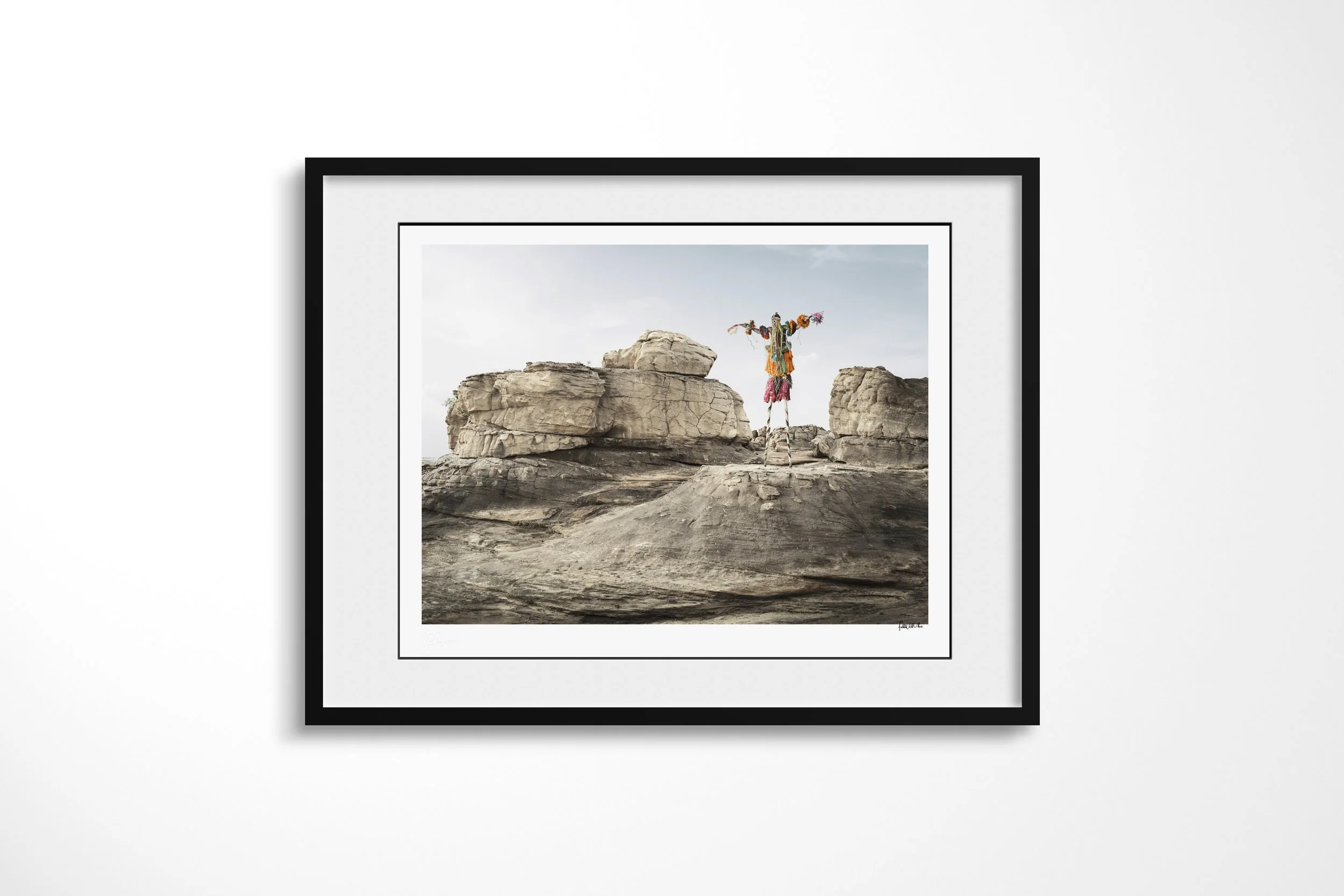 Framed presentation of Tegué Tanga (Mask) in a black frame and white mount, showing a Dogon stilt mask standing between broad sandstone outcrops.