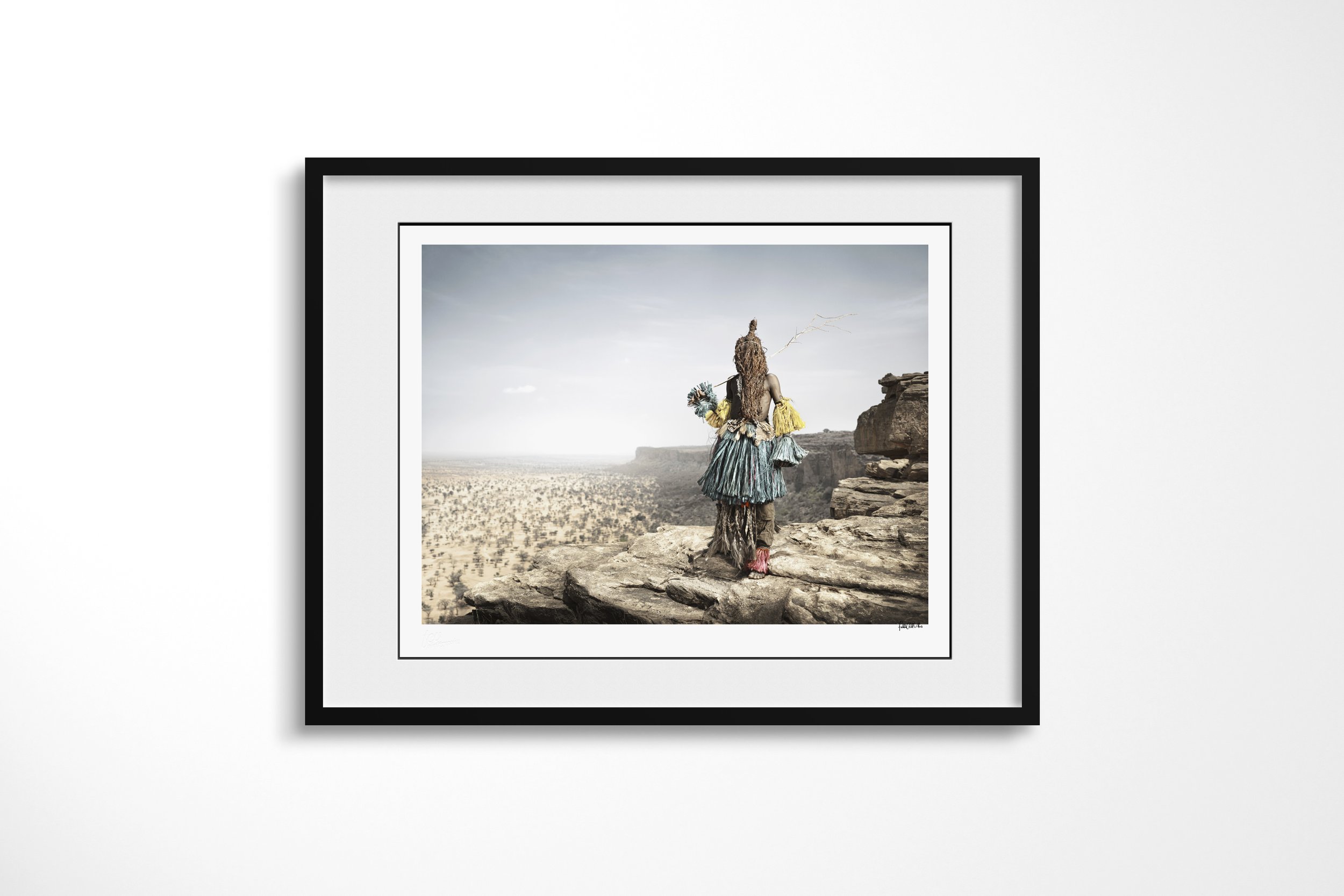Framed presentation of Benin Banin (Mask) in a black frame and white mount, showing a Dogon masked dancer seen from behind on a cliff overlooking the Bandiagara landscape.