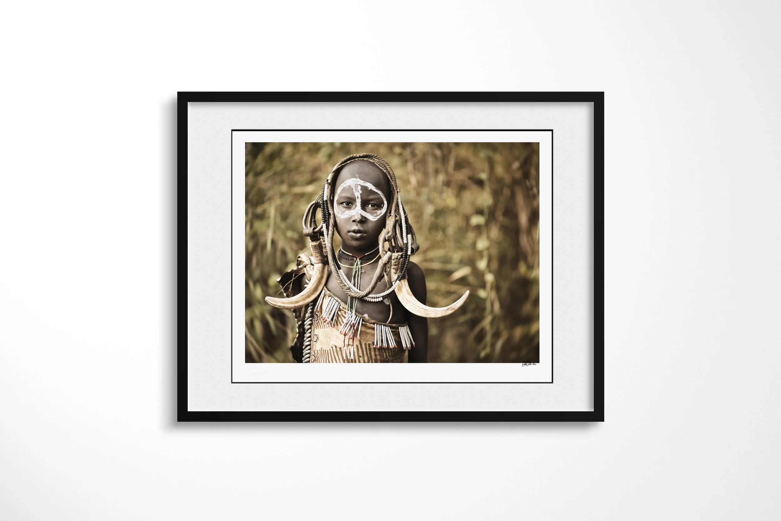 Framed presentation of Ngateri in a black frame and white mount, showing a young Mursi girl with white facial paint, beadwork and paired tusk ornaments against woodland.