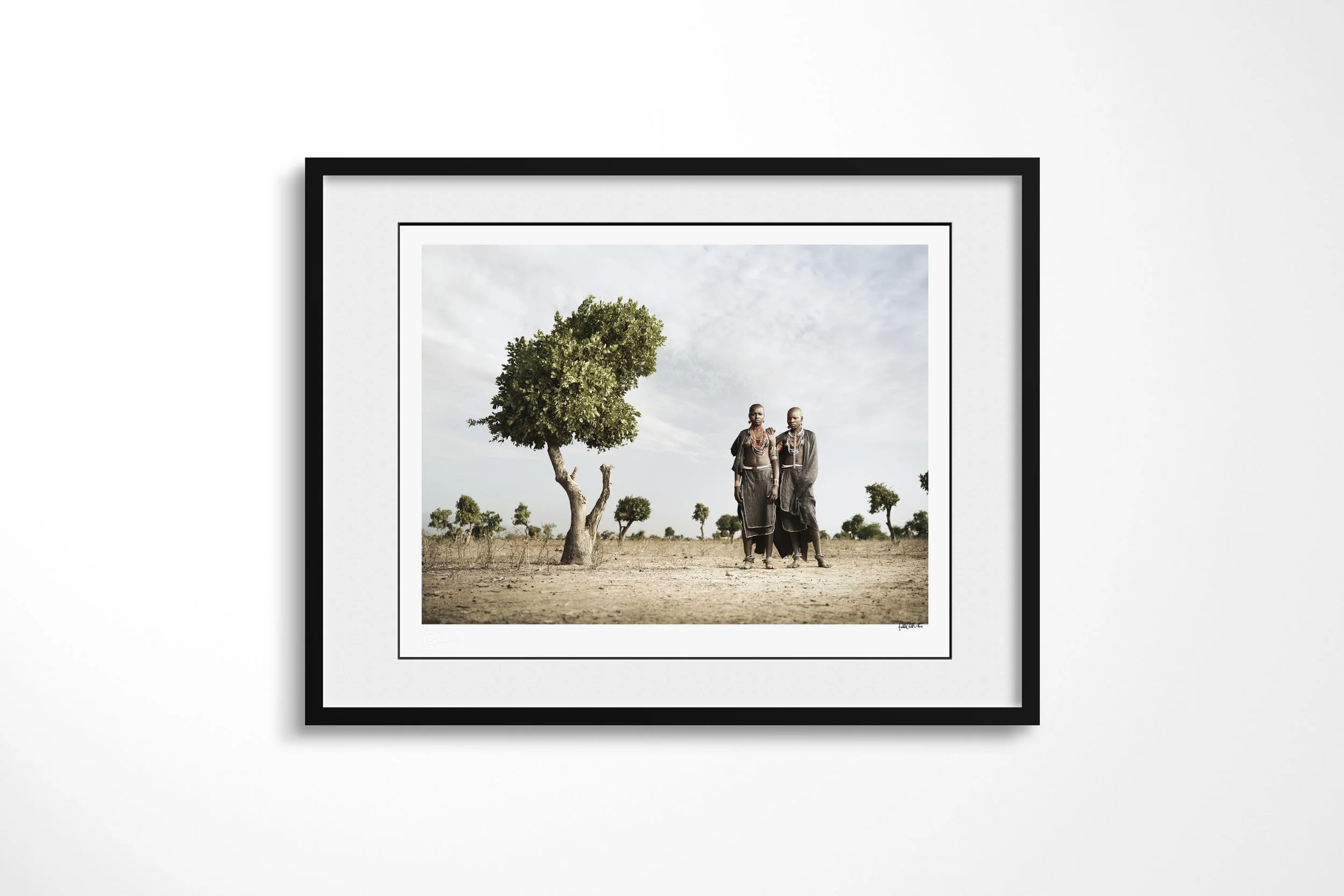 Framed presentation of Rufa & Lago in a black frame and white mount, showing two Arbore women standing together beneath a solitary tree in an arid landscape.