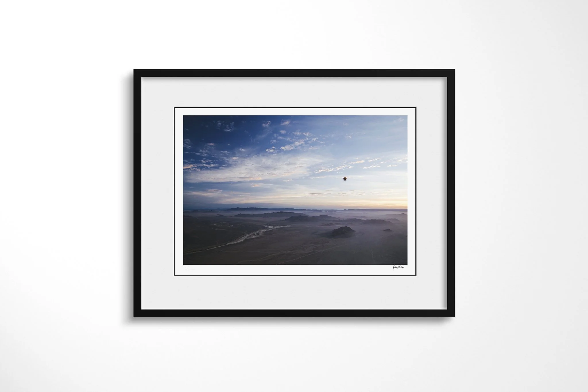 Framed presentation of Over The Dunes in a black frame and white mount, showing small hot-air balloon above an expansive arid landscape of ridges, valleys and pale winding channels at dawn.