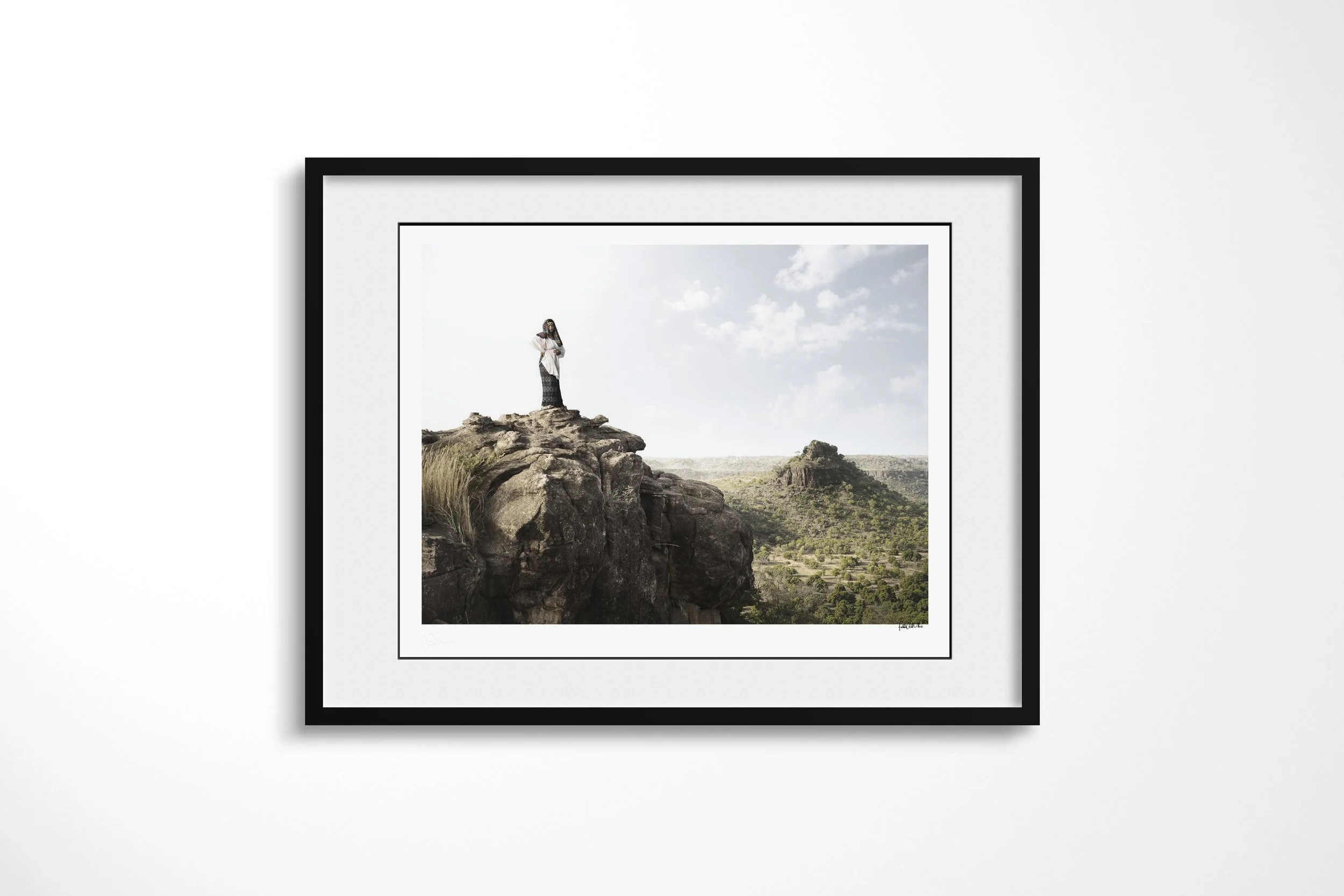 Framed presentation of Fante in a black frame and white mount, showing Fante standing on a high rocky ledge above a green valley and distant escarpment.