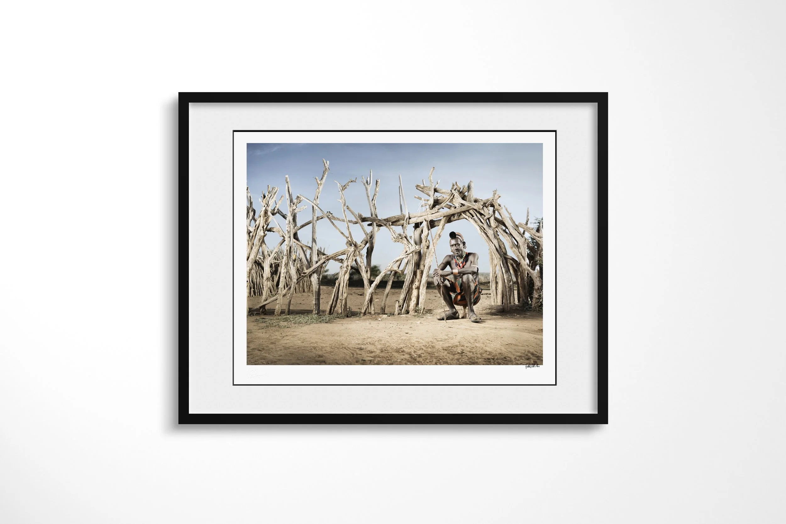 Framed presentation of Woro in a black frame and white mount, showing an older Hamar man with pale body paint and layered beadwork crouching inside a branch-built enclosure.