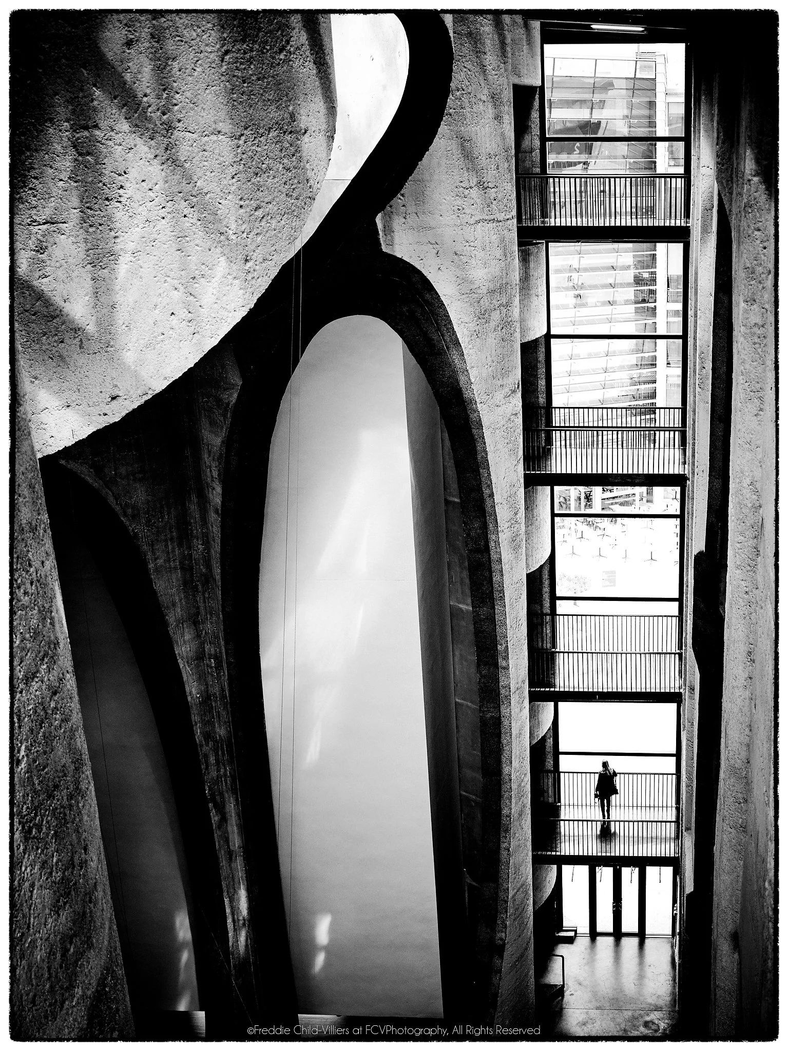 Monumental curved concrete forms beside stacked walkways, with a solitary figure visible in a high-contrast interior.