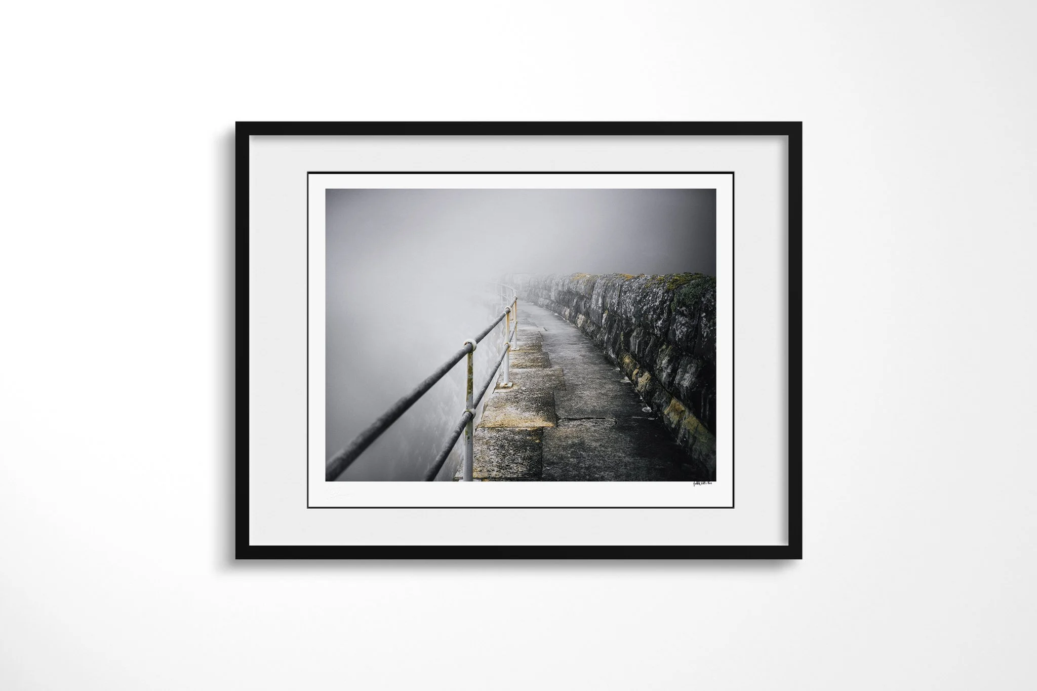 Framed fine art presentation of Ballustrade in a black frame with a white mount, depicting a weathered concrete walkway between a metal handrail and stone wall, curving out of sight into dense fog.