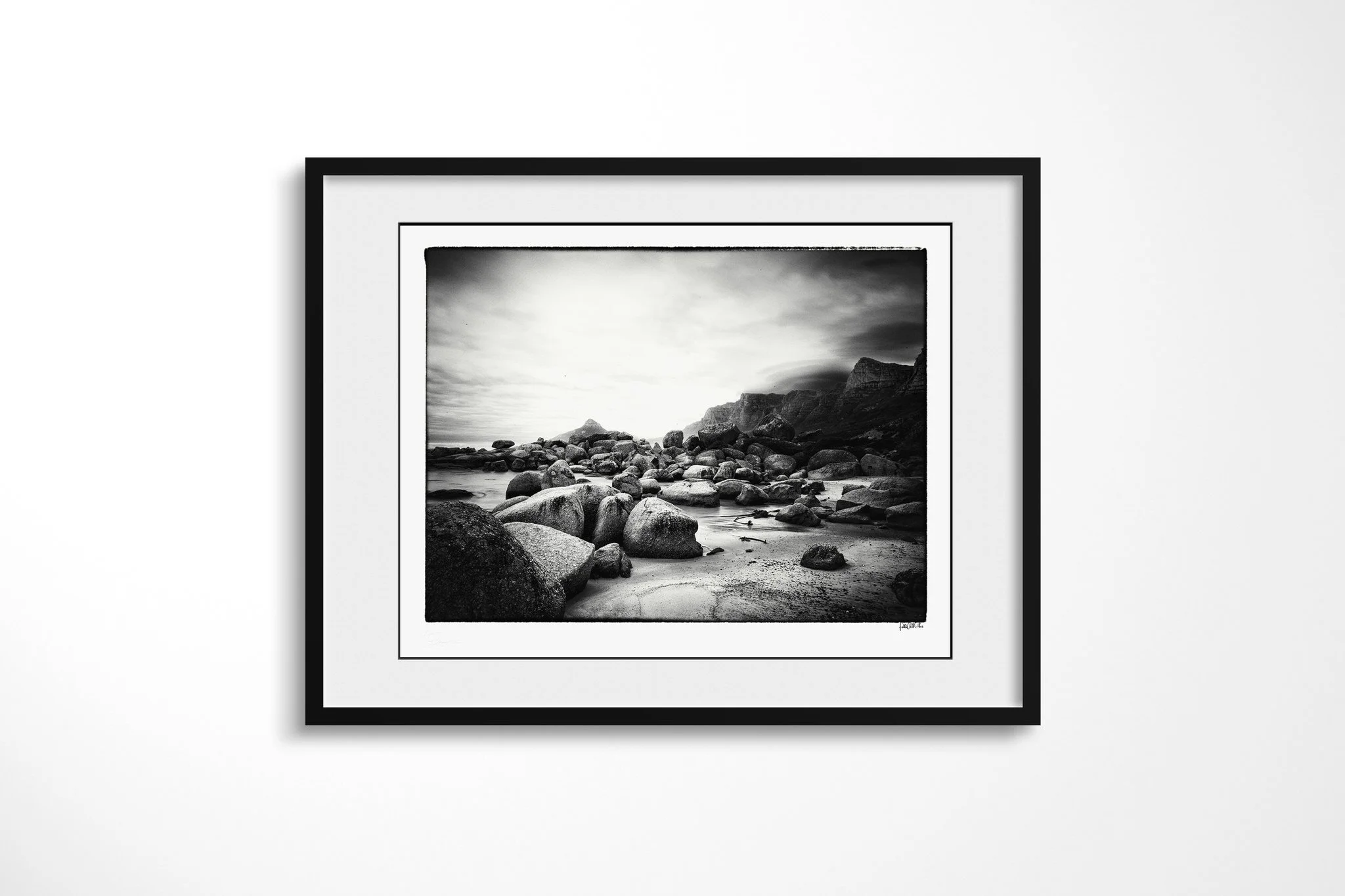 Twelve Apostles presented in a black frame with a white mount, depicting a black-and-white boulder beach beneath Cape Town's Twelve Apostles cliffs, with low cloud and softened seawater.