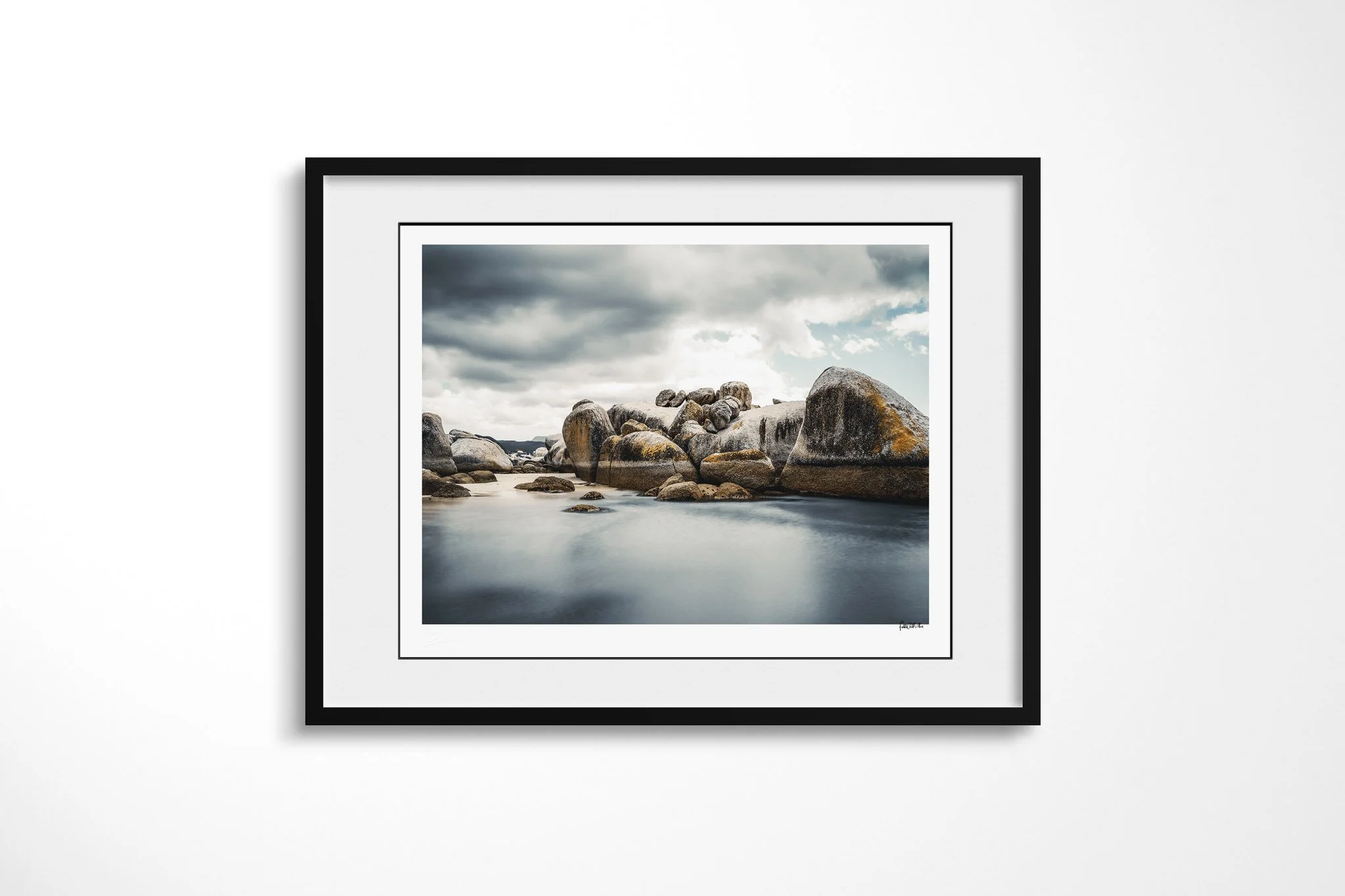 Framed presentation of Tideline in a black frame and white mount, showing rounded granite boulders around smooth blue-grey coastal water beneath heavy clouds, with patches of warm lichen.