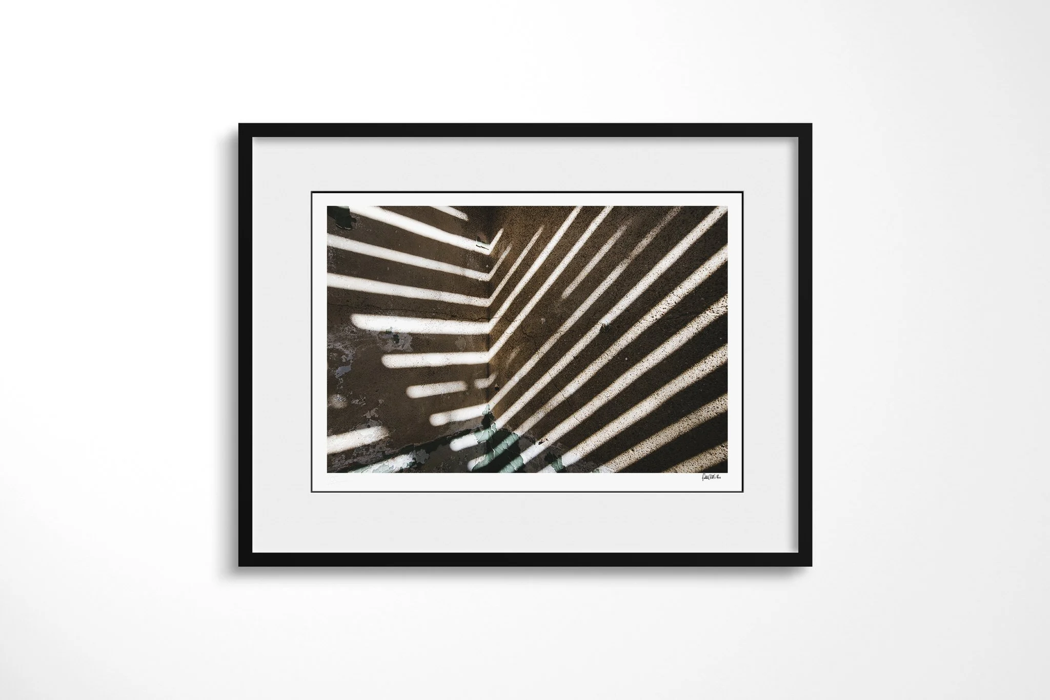 Black-framed fine art print of Streak with a white mount, showing diagonal bands of sunlight and shadow crossing a damaged interior, with cracked surfaces and a small green plant below.