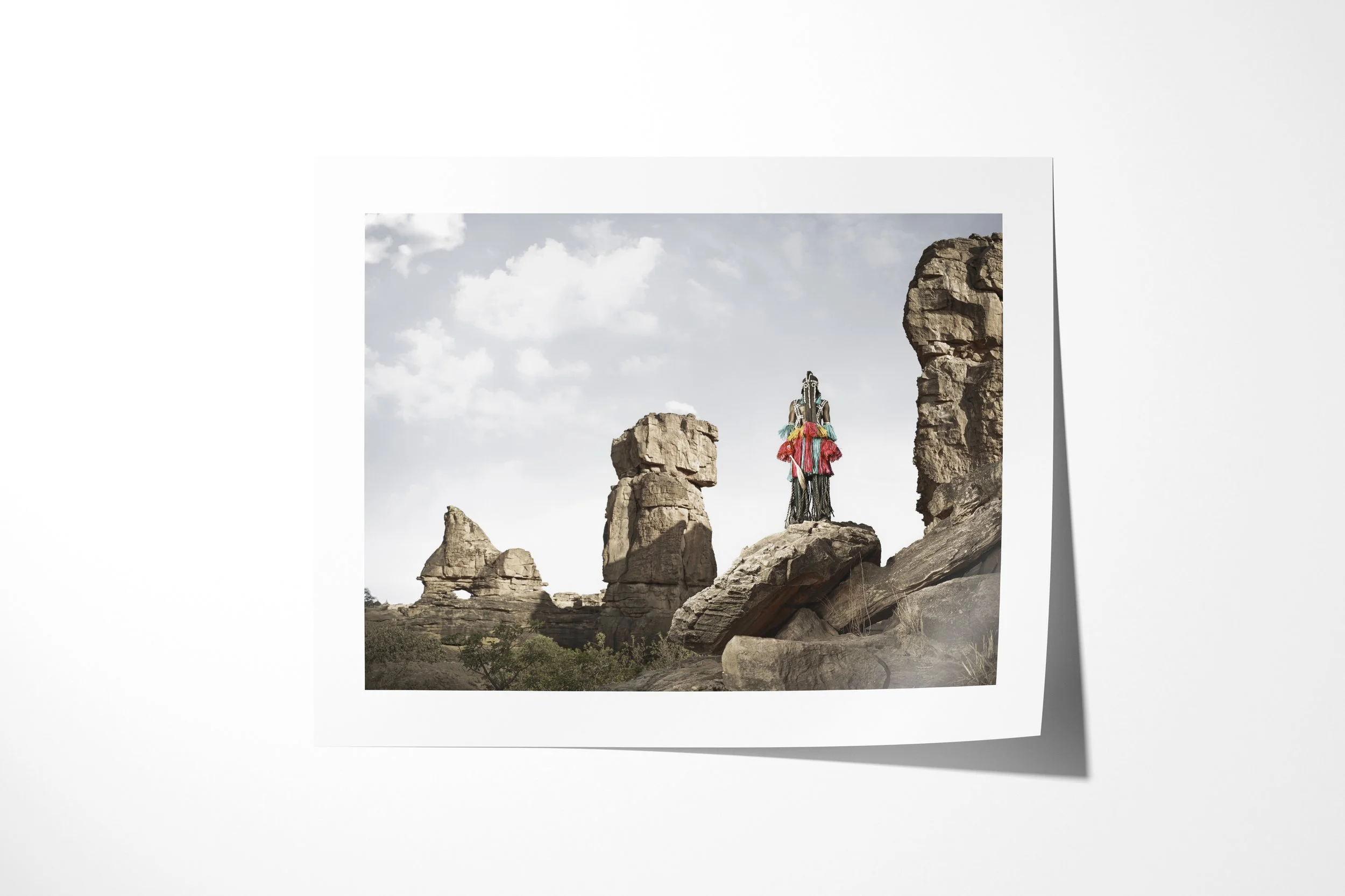 Fine art paper print of Lagassa Lyai (Mask), showing a Dogon masked figure standing high among monumental sandstone formations.