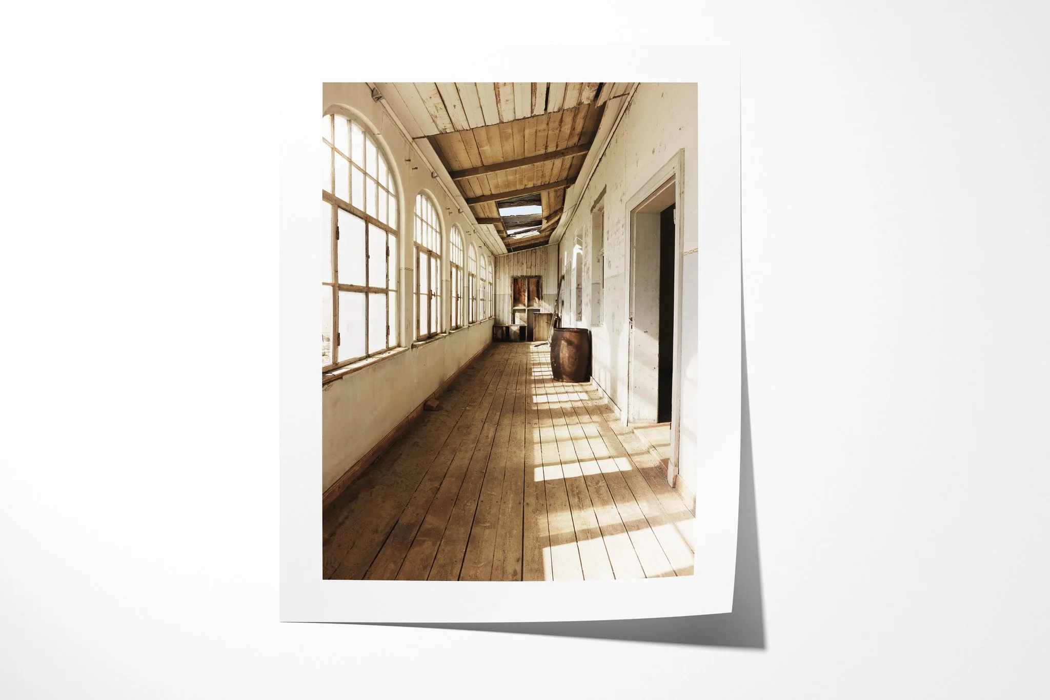 Fine art paper print of School Corridor, showing a long abandoned school corridor with arched windows, worn wooden floorboards, open doors and sunlight crossing peeling walls.