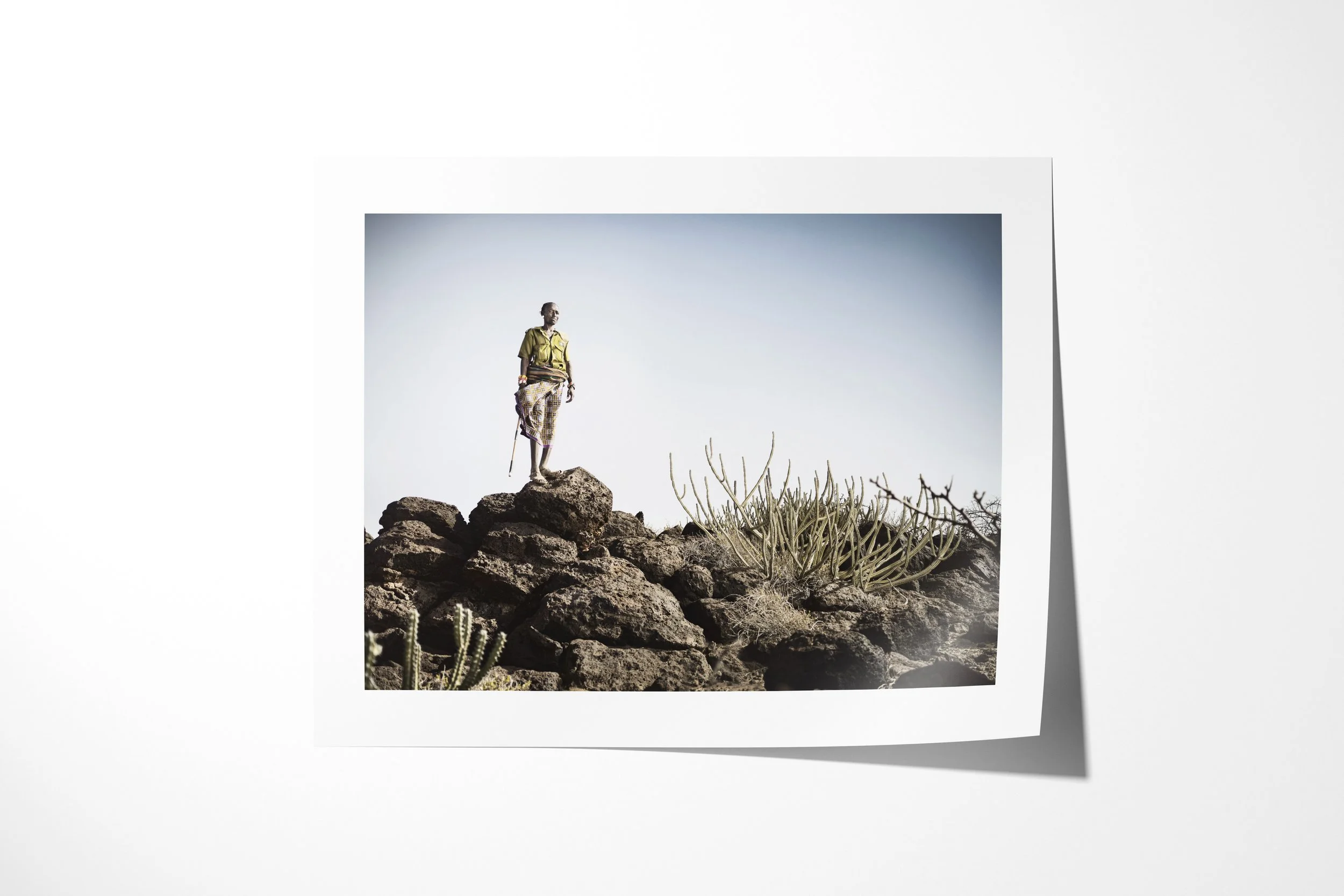 Fine art paper print of Lpushe - “The Shepherd”, showing a Rendille herder holding a staff on black rocks beside a desert succulent.