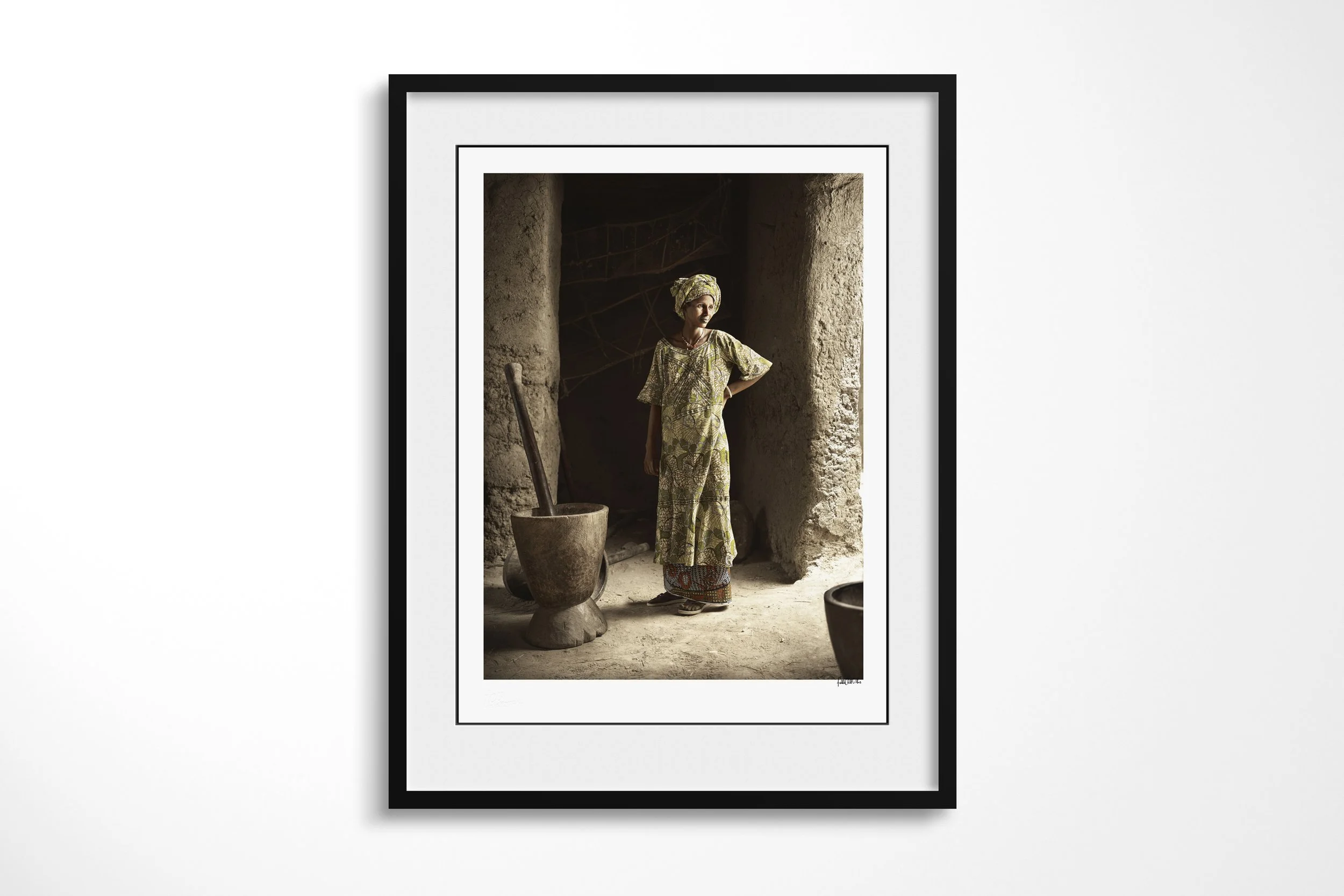 Framed presentation of Tiedo in a black frame and white mount, showing Tiedo standing beside a large wooden mortar in the doorway of a dim earthen building.