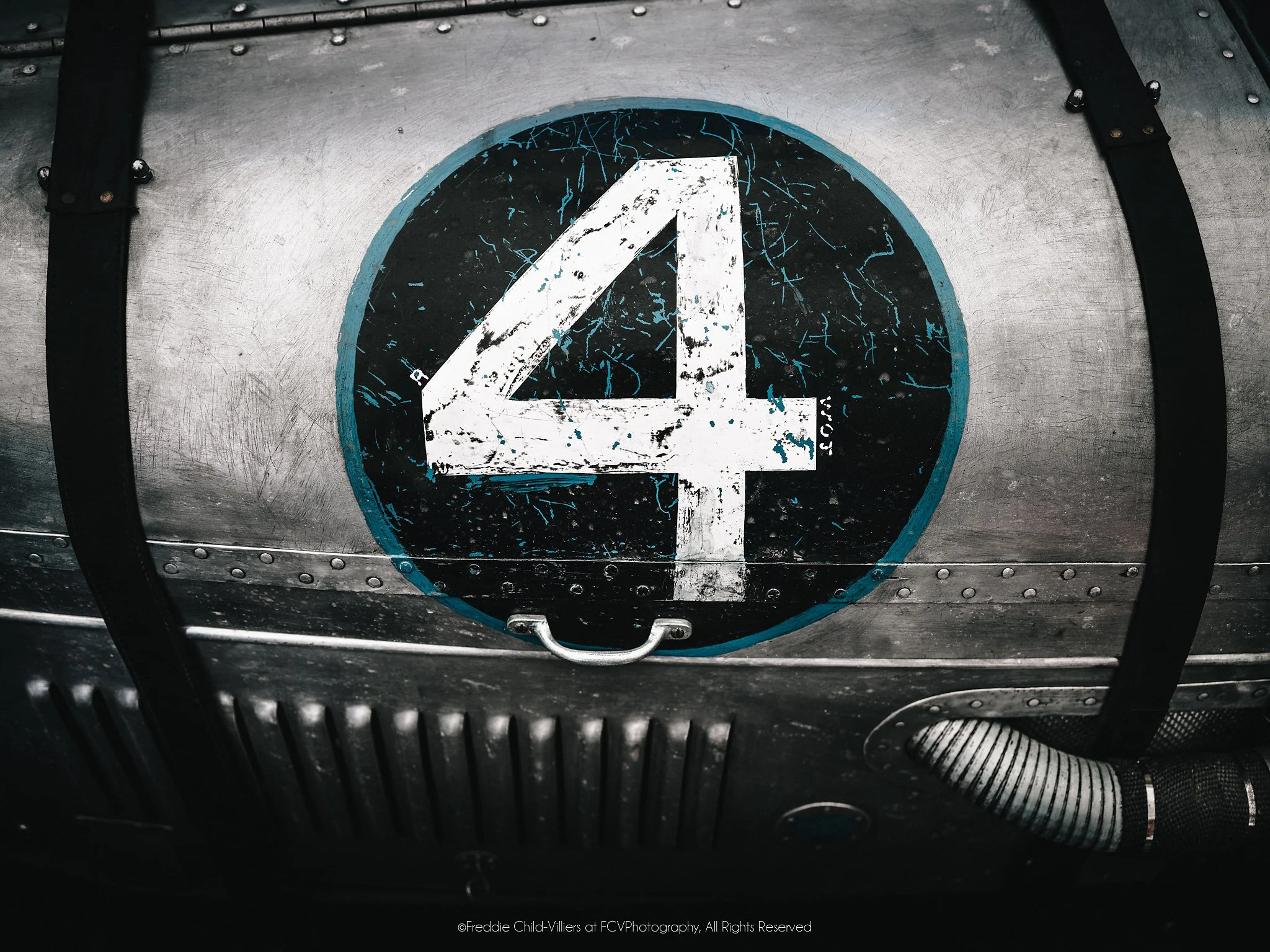 Weathered white numeral four on a black circular plate set into riveted metal with straps and ribbed tubing.