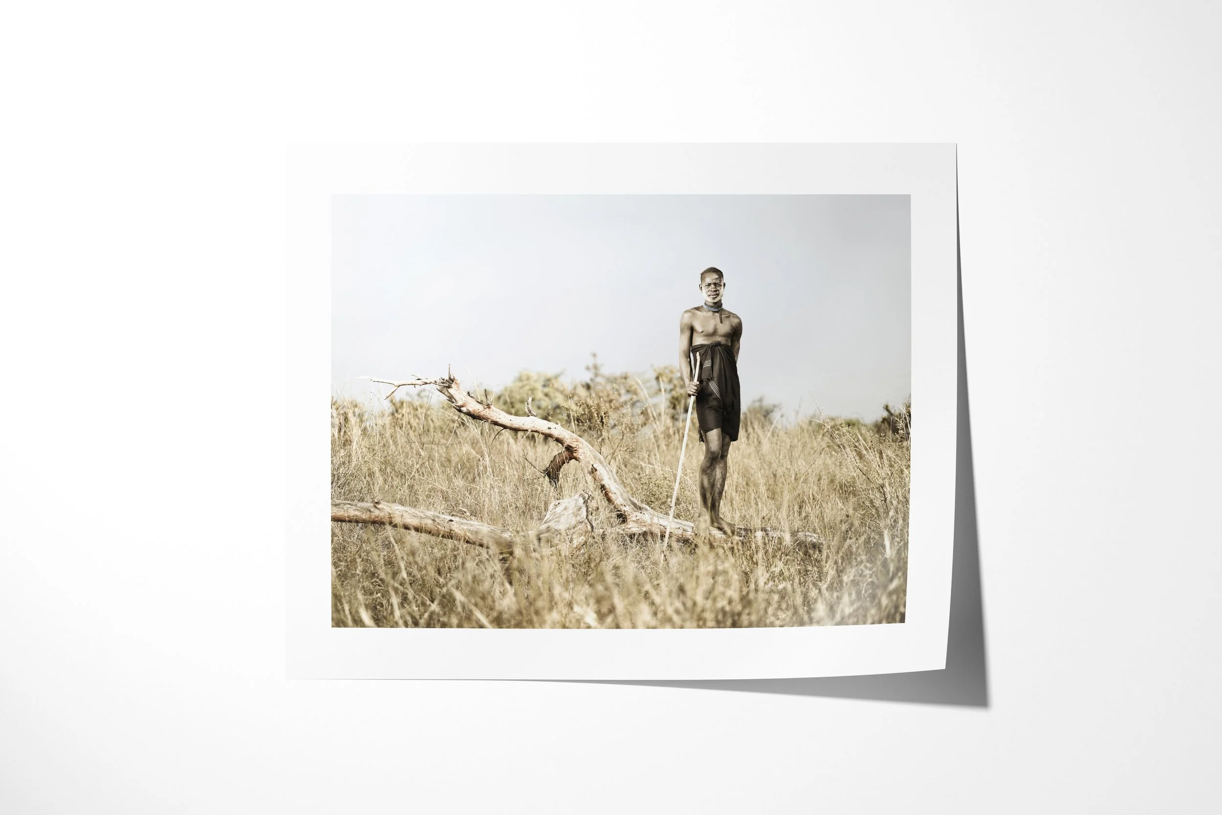 Fine art paper print of Morkoro, showing a Mursi man in a dark wrap standing with a staff beside a fallen branch in dry grassland.
