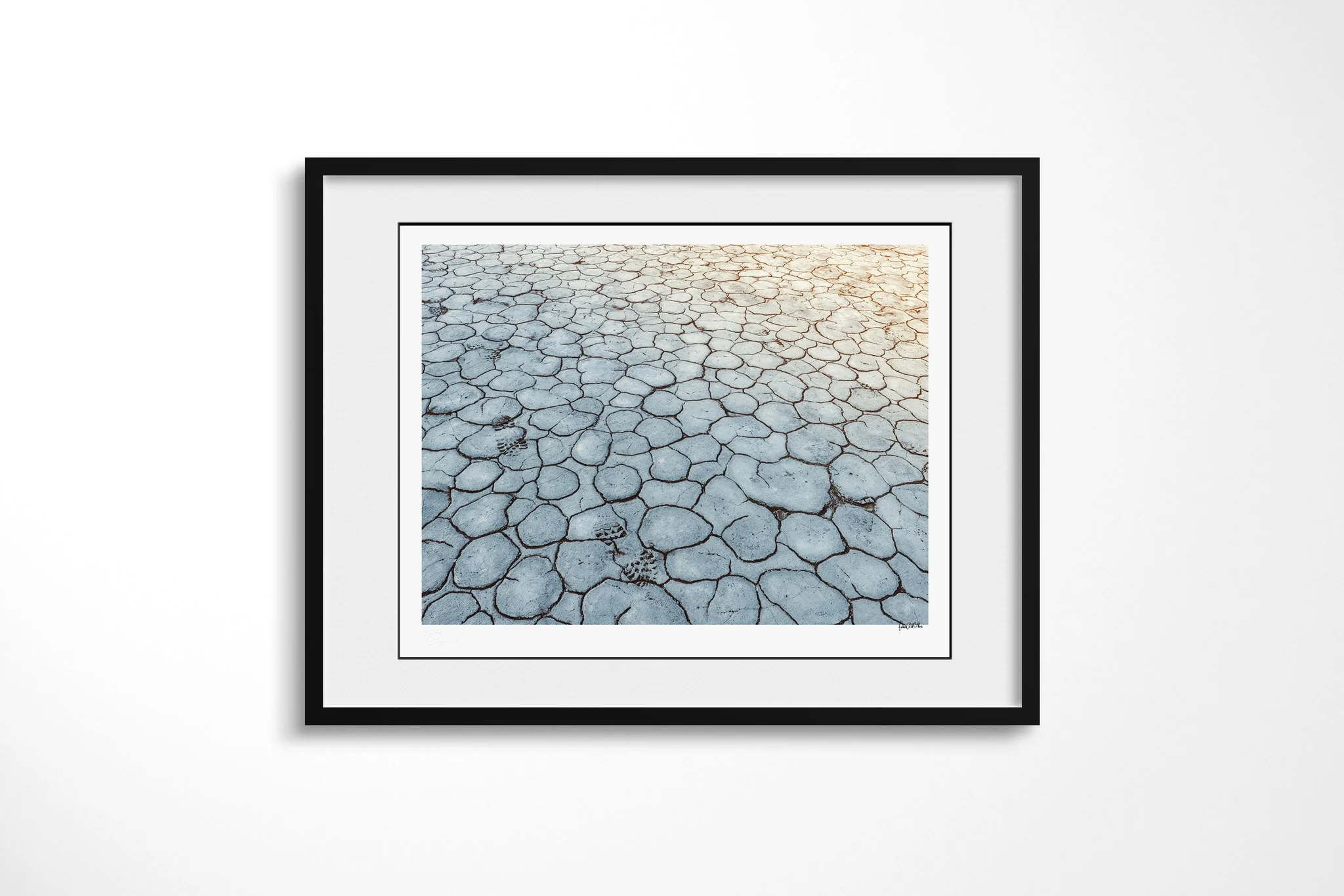 Footsteps presented in a black frame with a white mount, depicting a trail of shoeprints crossing pale blue-grey cracked earth, with warm light entering from the upper right.