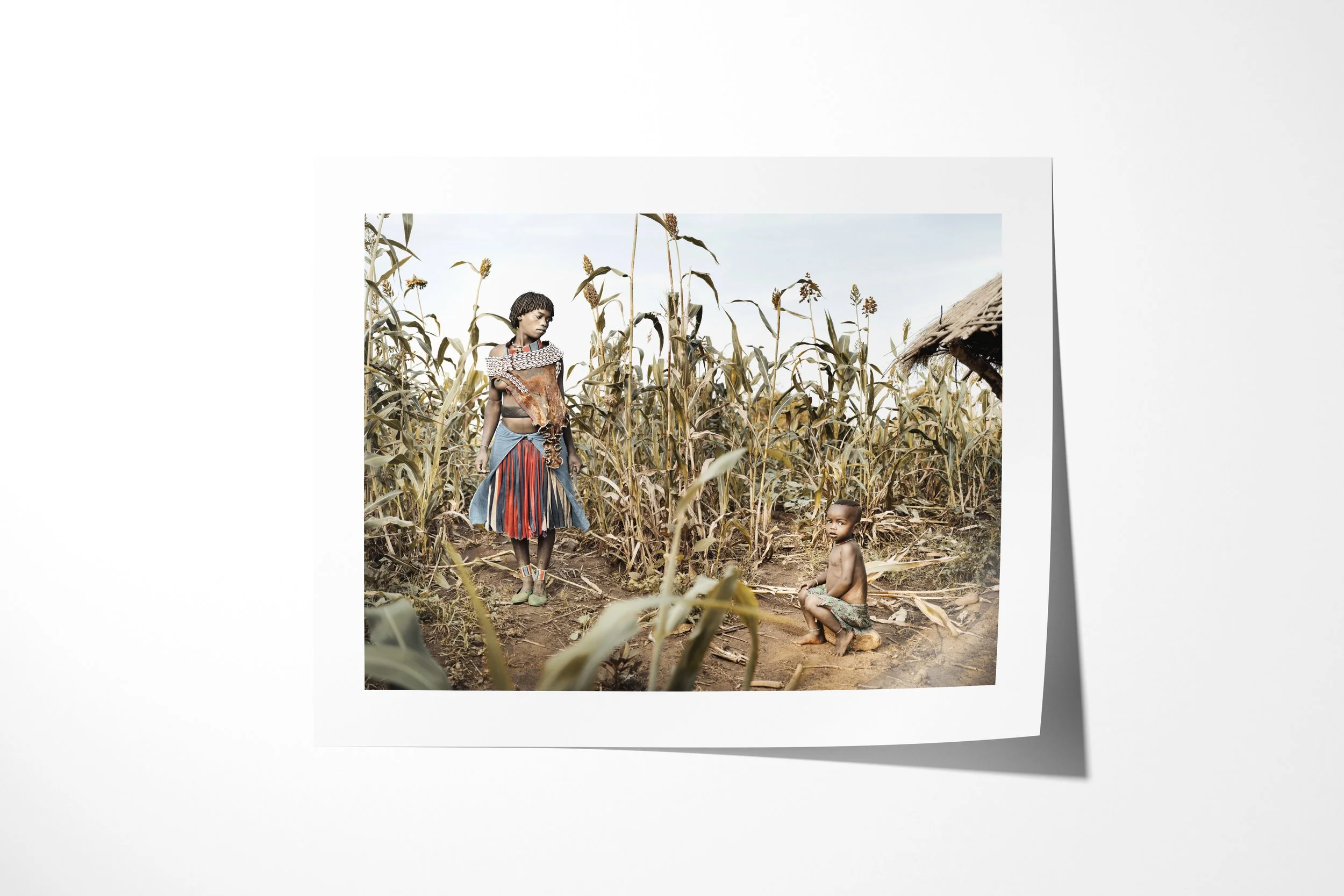 Fine art paper print of Haila & Wancho, showing a Banna woman and child standing among tall sorghum beside a thatched structure.