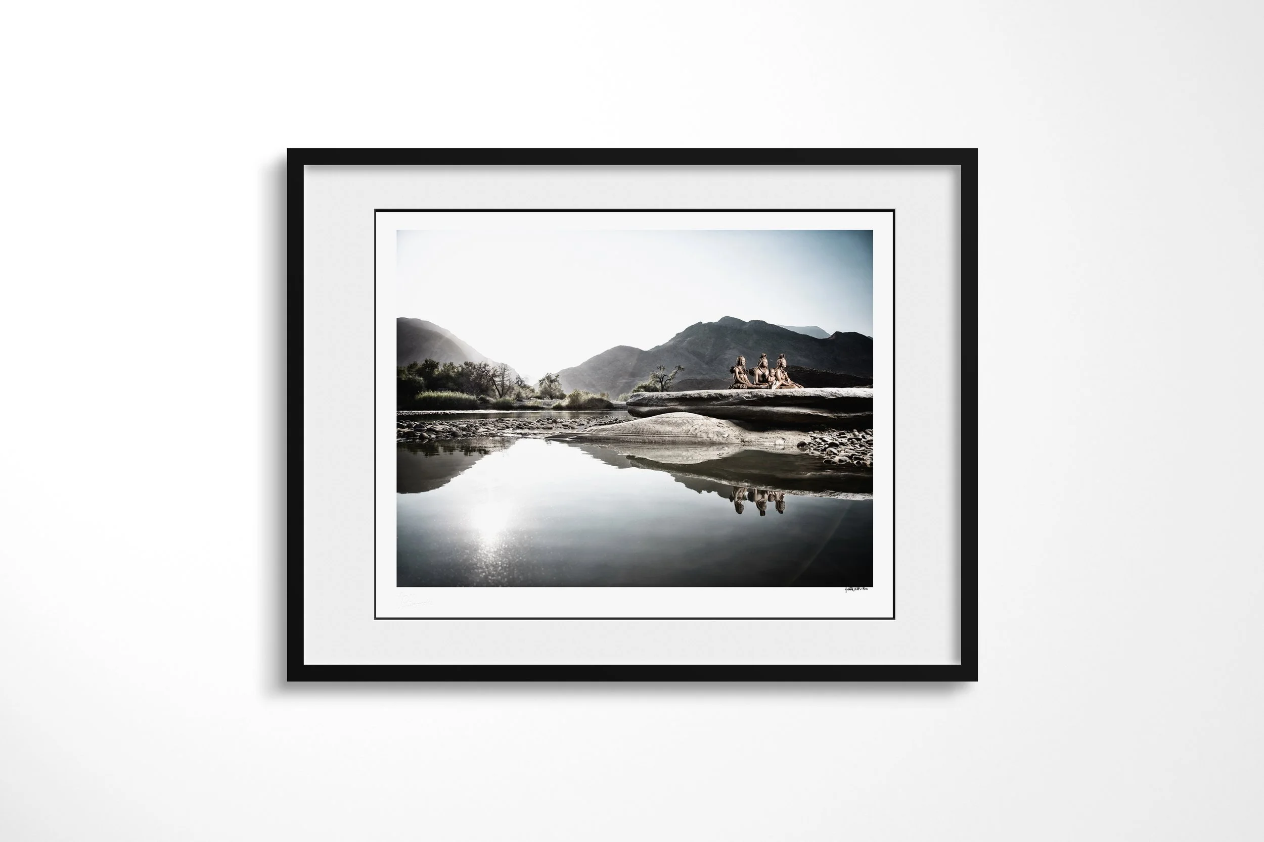 Framed presentation of Mbendura, Tjikuria and Mutume in a black frame and white mount, showing a Himba family gathered on a broad rock beside the Kunene River with their reflections in the water.