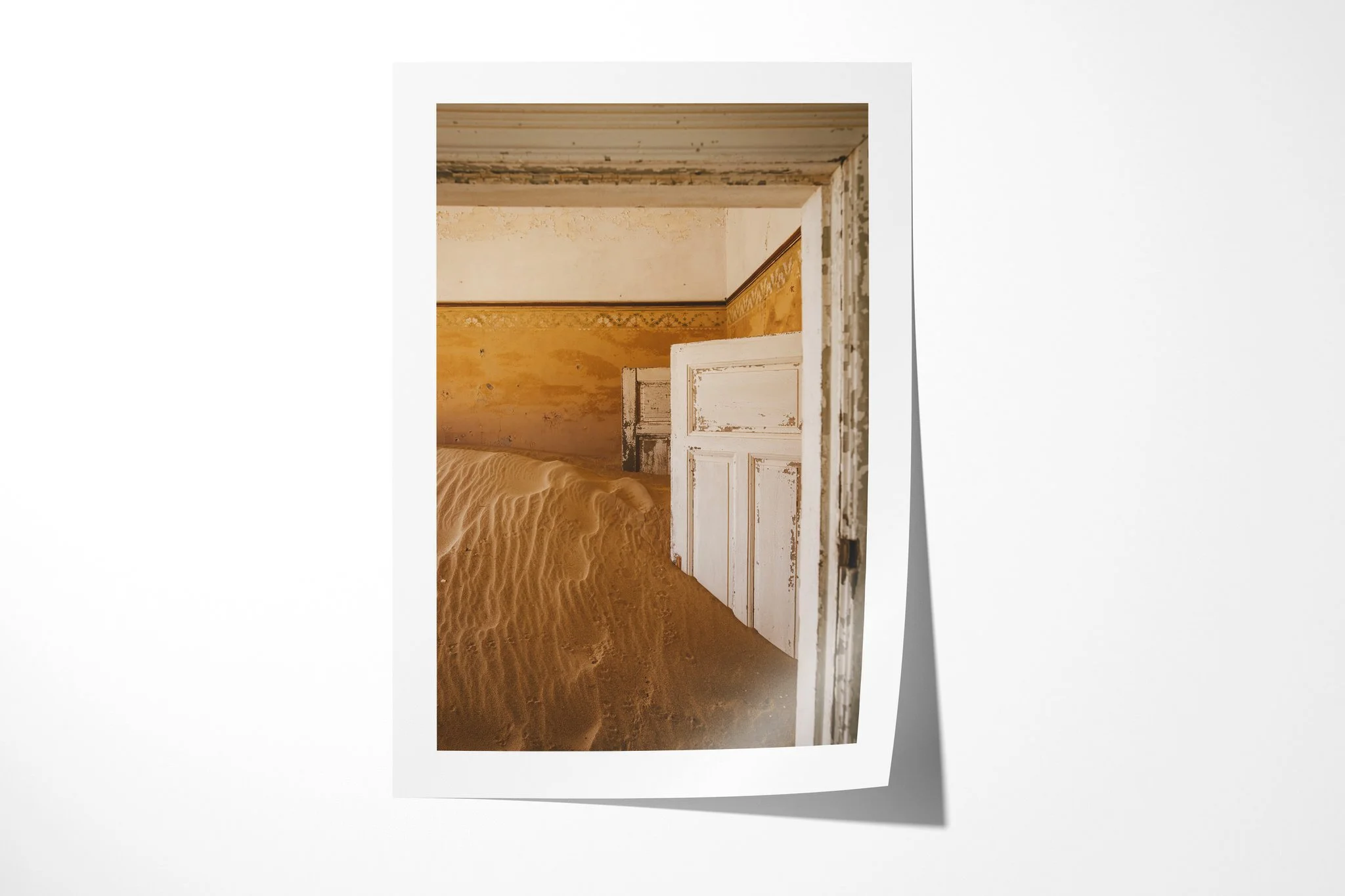 Unframed fine art paper print of Doorway III, showing chipped white doors and ochre walls layered behind a high drift of rippled sand in an abandoned interior.