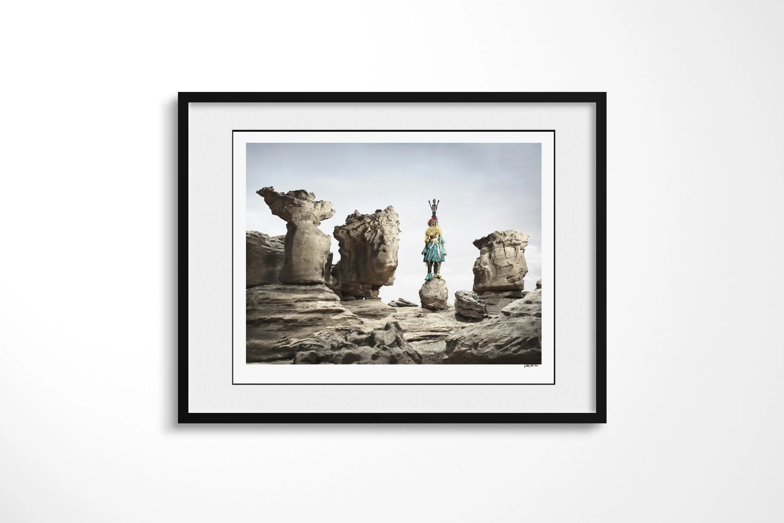 Framed presentation of Satimbe (Mask) in a black frame and white mount, showing a Dogon Satimbe mask balanced on a small rock among weathered stone pillars.