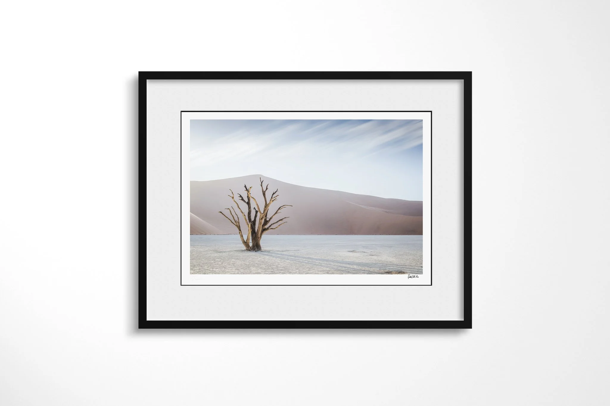Black-framed fine art print of Deadvlei with a white mount, showing a dead tree on the pale Deadvlei clay pan before a vast desert dune under streaked clouds.
