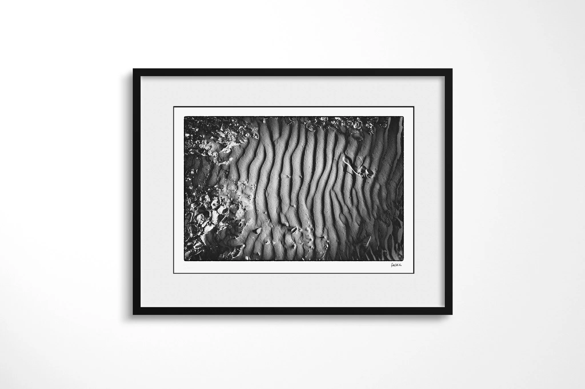 Framed presentation of Khamsin VII in a black frame with a wide white mount, showing a vertical band of sand ripples bordered by broken crust, with a small curled plant fragment.