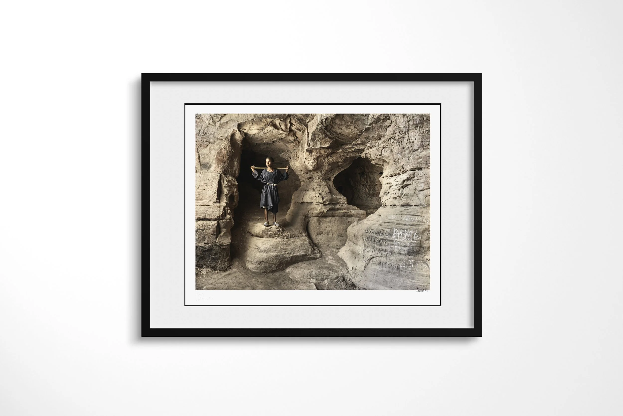 Framed presentation of Modibo in a black frame and white mount, showing Modibo standing with outstretched arms inside a cave marked with handwritten requests.