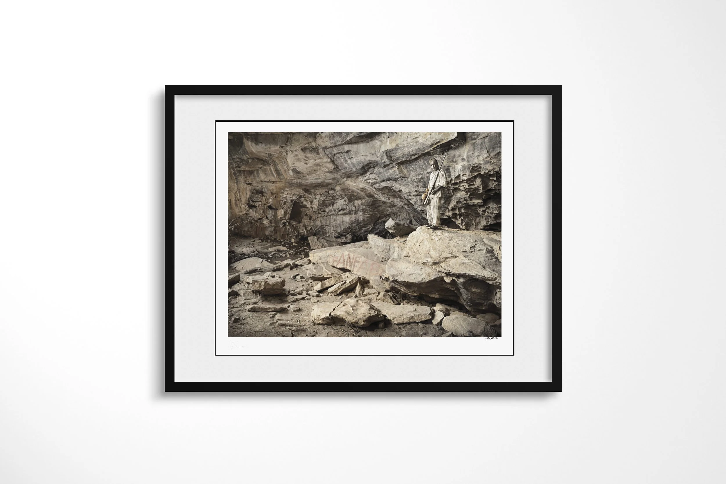 Framed presentation of Bomary in a black frame and white mount, showing Bomary standing on a rock ledge within the broad chamber of a sacred cave.