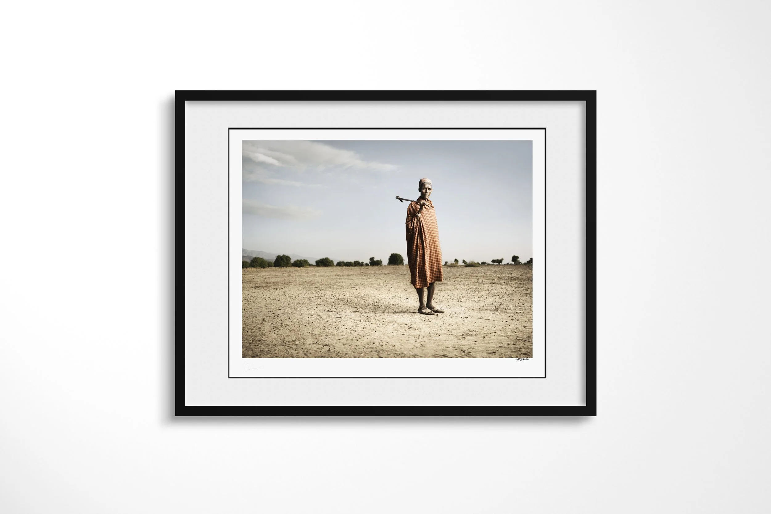 Framed presentation of Aregore in a black frame and white mount, showing an older Arbore man wrapped in checked cloth standing with a staff across his shoulders on open ground.
