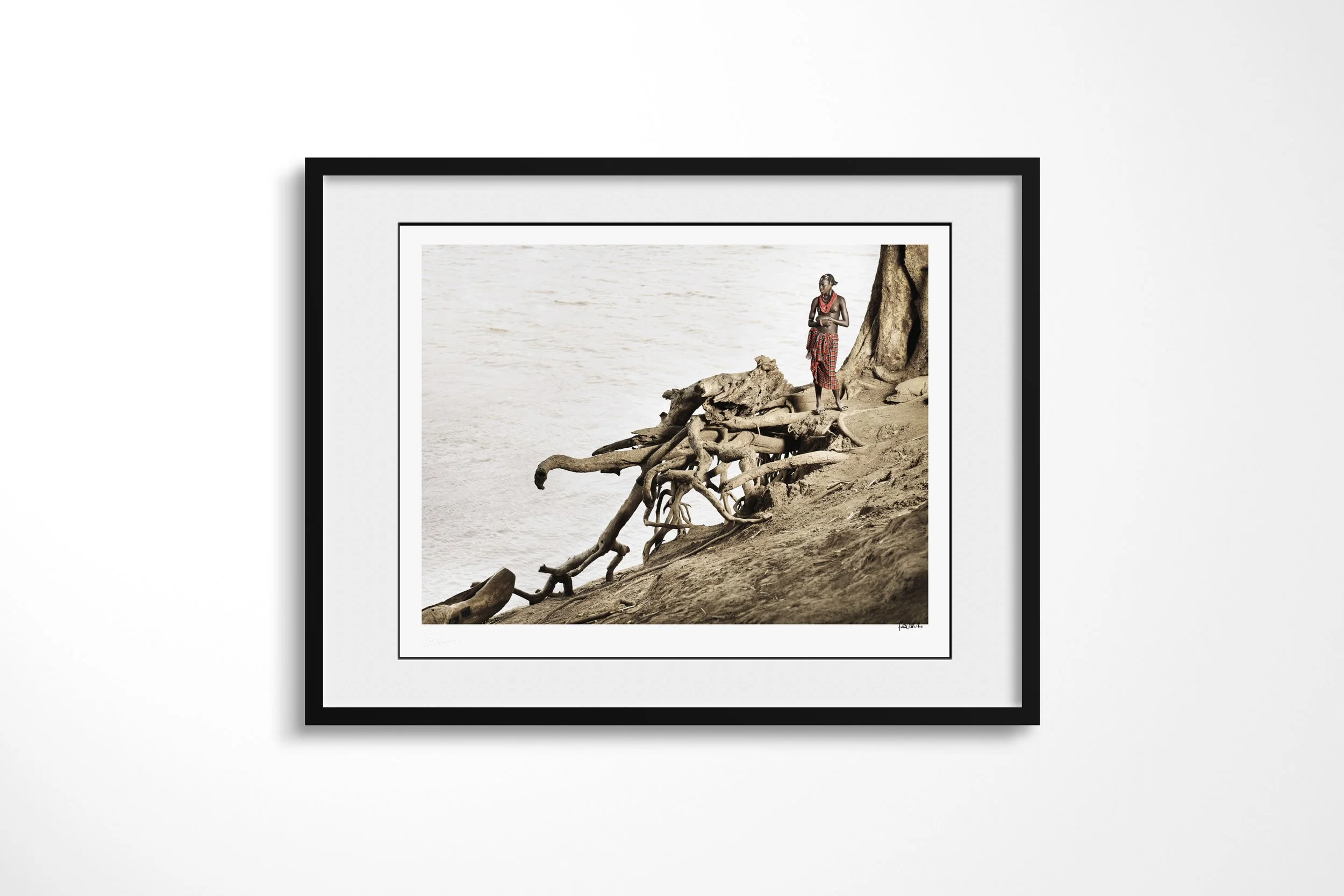Framed presentation of Yierar in a black frame and white mount, showing a Daasanach man standing beside exposed tree roots on the bank of the Omo River.