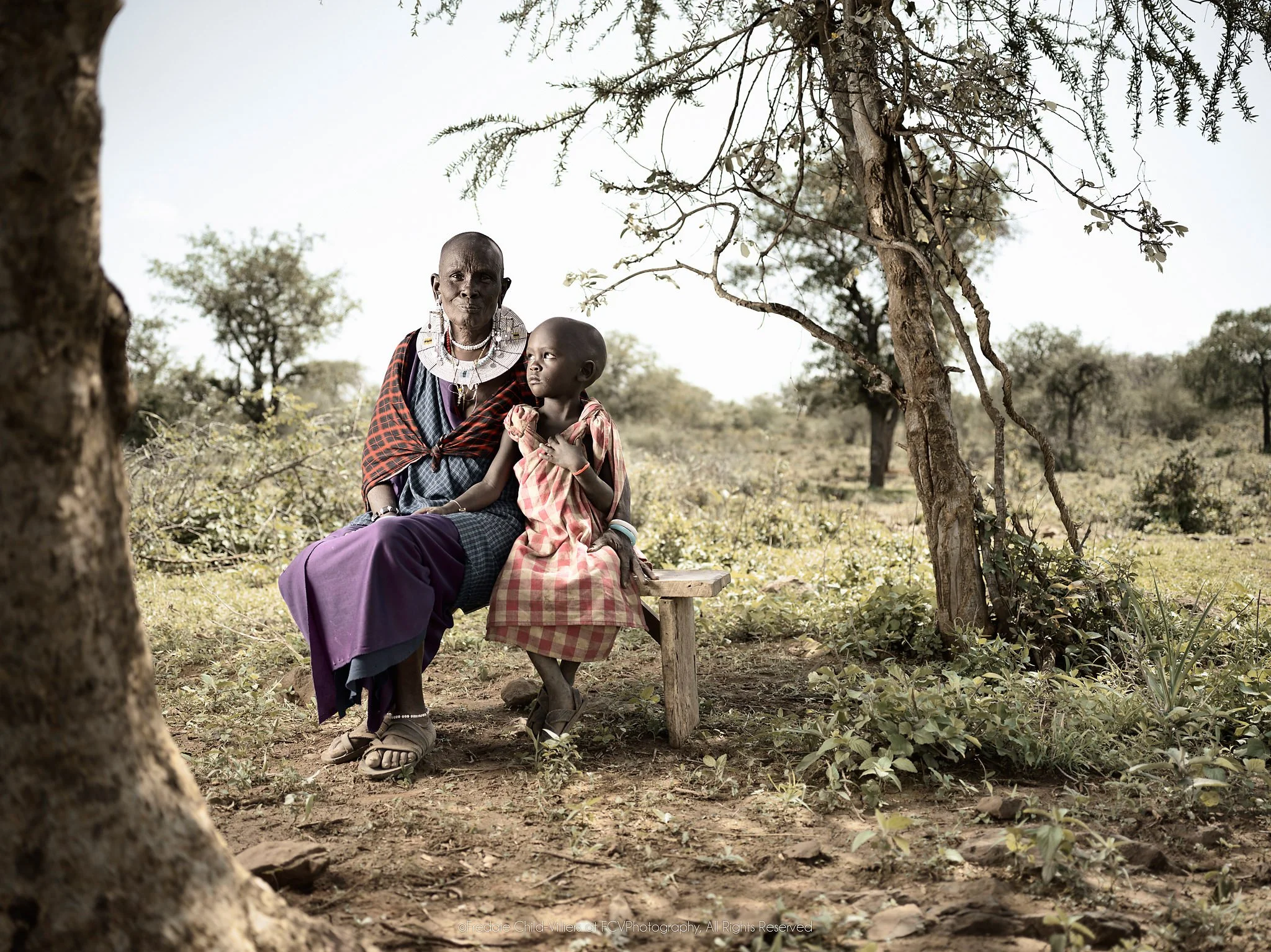 Naleku seated beneath trees with her young grandson Elias standing beside her.