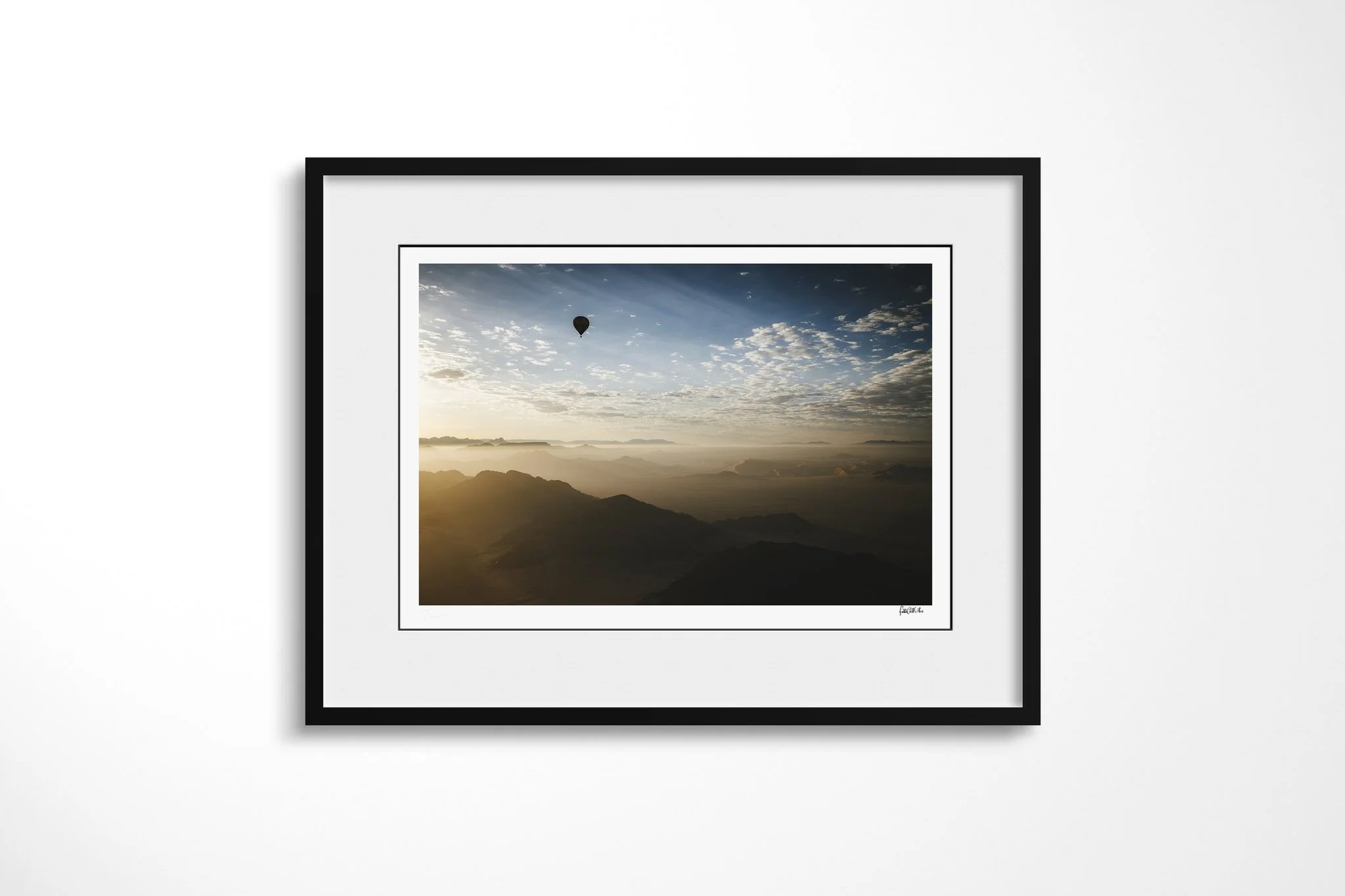 Framed presentation of 80 Days in a black frame and white mount, showing a lone hot-air balloon above layered arid mountain ridges in warm early light beneath scattered clouds.