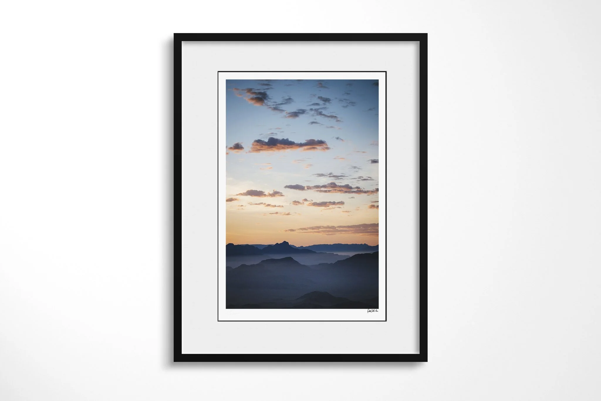Framed presentation of Sossusvlei Sunrise in a black frame and white mount, showing layered dark desert ridges at Sossusvlei beneath peach and pale blue clouds at sunrise.