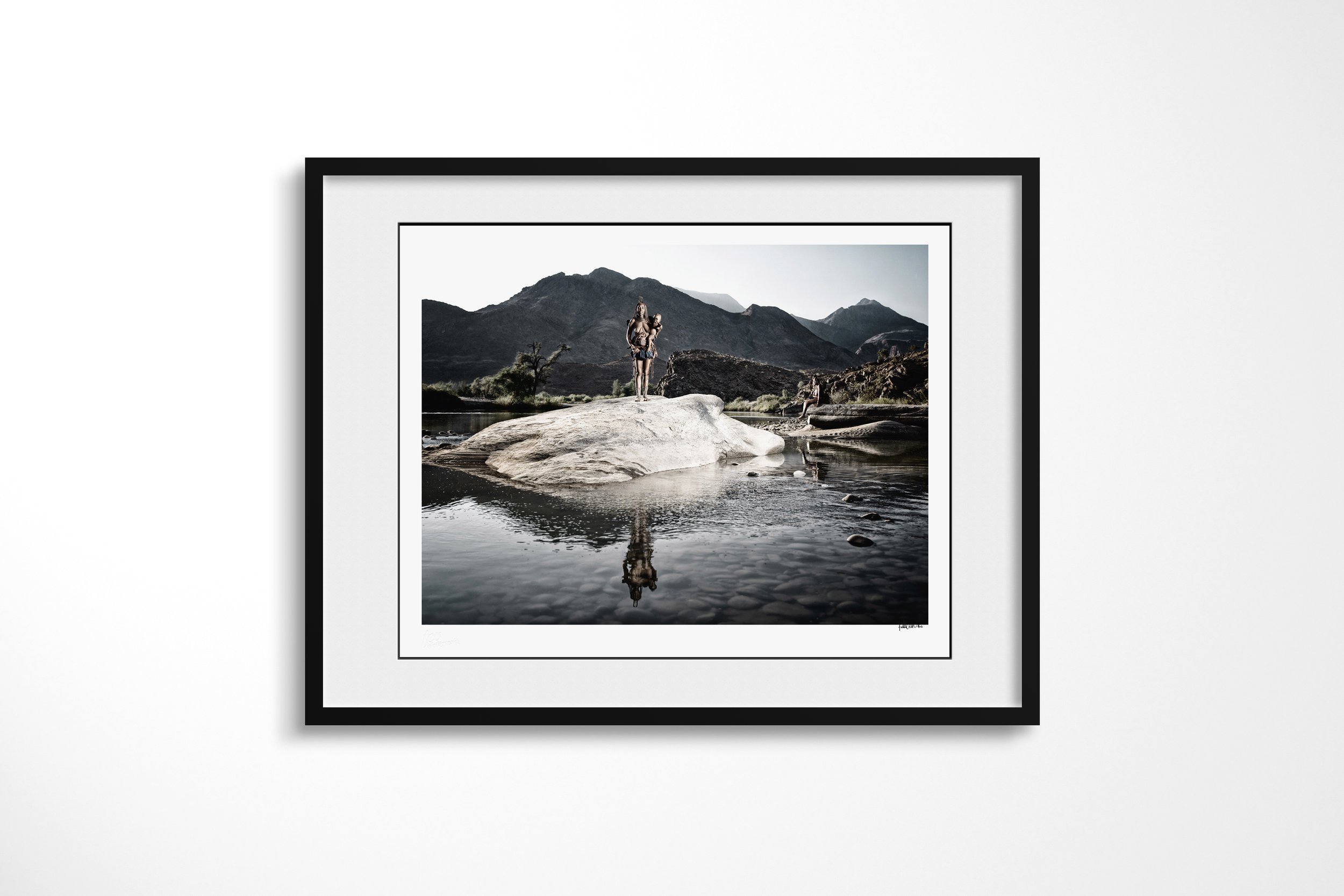 Framed presentation of Tunae (foreground), Katafa (background) in a black frame and white mount, showing two distant figures standing on a river boulder, reflected in the still water below.