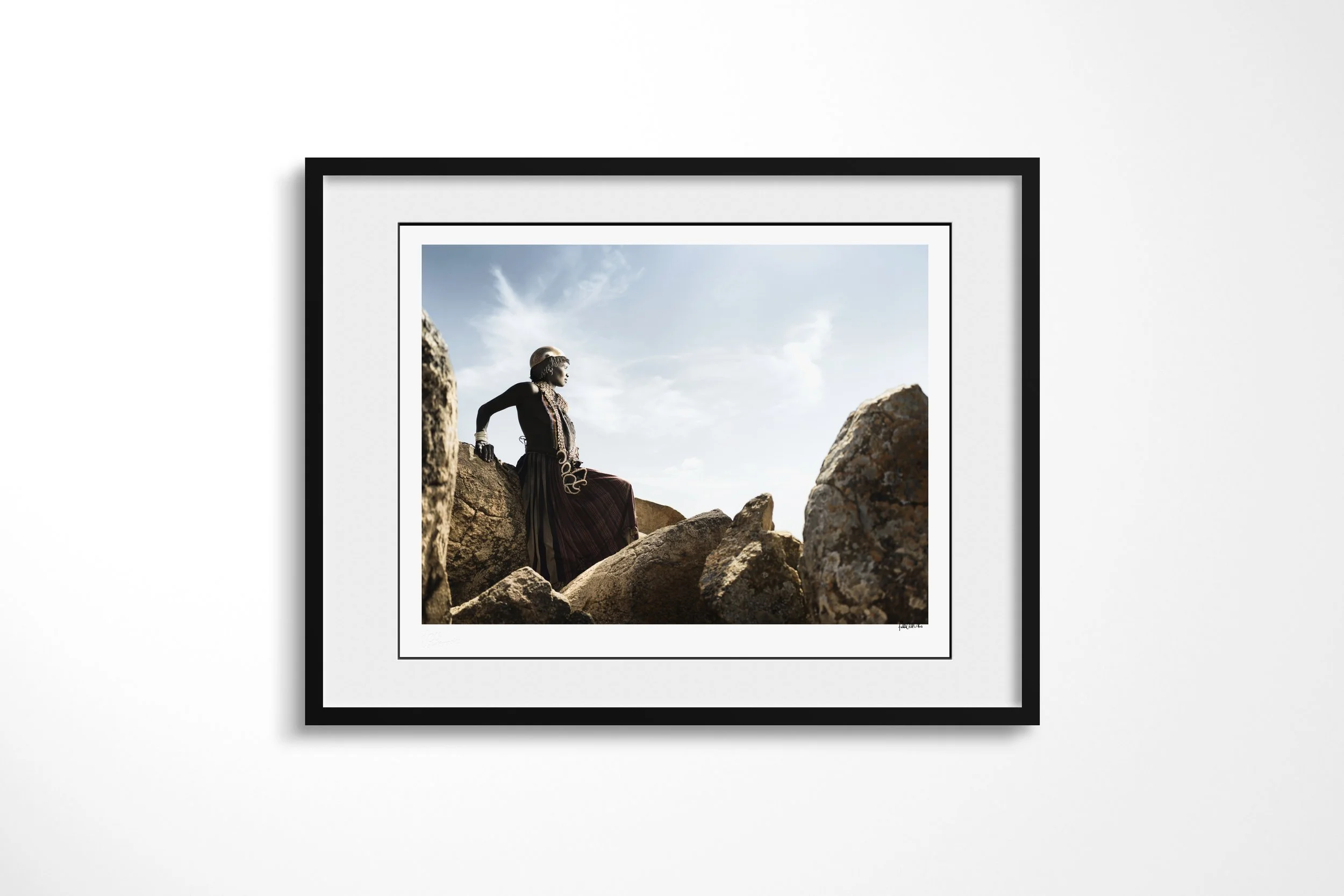 Framed presentation of Koiga in a black frame and white mount, showing a Banna woman in a long dark dress sitting among large sunlit rocks beneath an open sky.