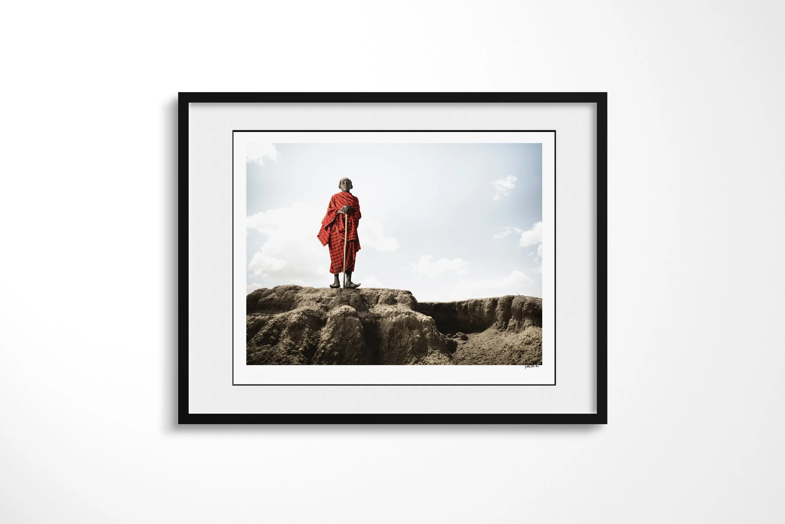 Framed presentation of Saitoti in a black frame and white mount, showing a Maasai boy in a red checked wrap standing with a herding staff on an eroded ridge.