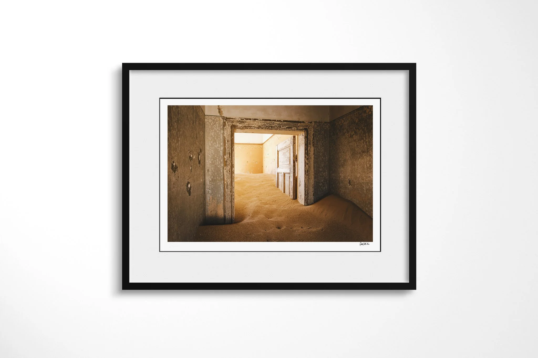 Doorway II presented in a black frame with a white mount, depicting open doorways leading through worn ochre rooms whose floors are covered by rippled desert sand.