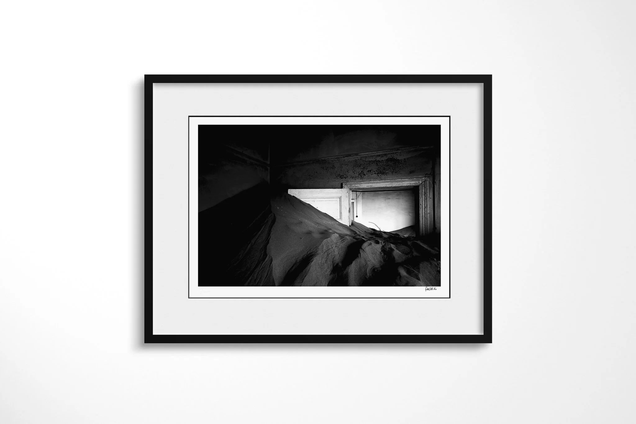 Framed presentation of Doorway I in a black frame and white mount, showing a steep sand drift filling a dark abandoned room and rising through a partly open doorway in black and white.